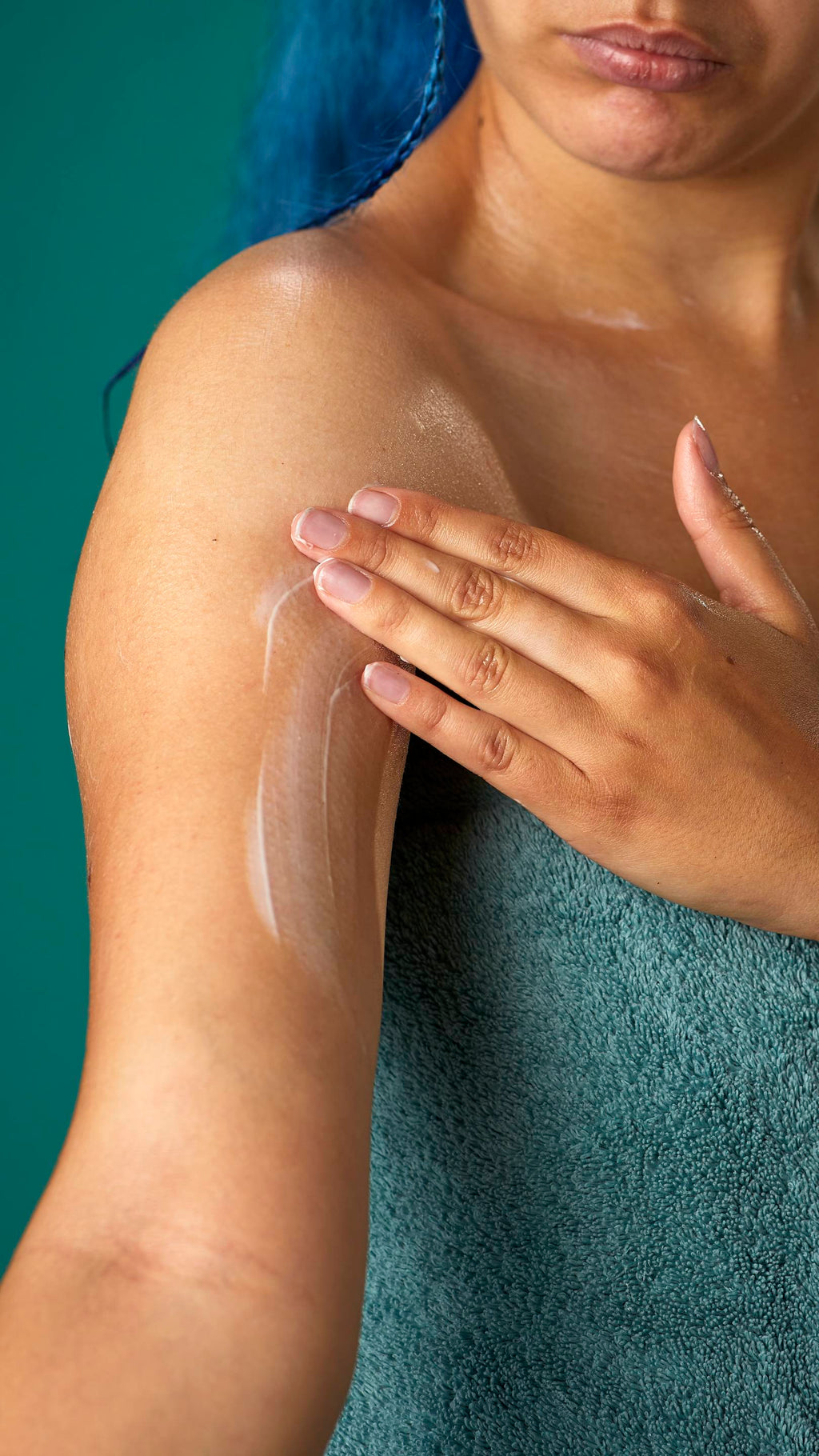 The image shows a close-up of the model, wrapped in a sage-green towel on a teal background. They are gently applying the Coconut body butter to their upper arm.