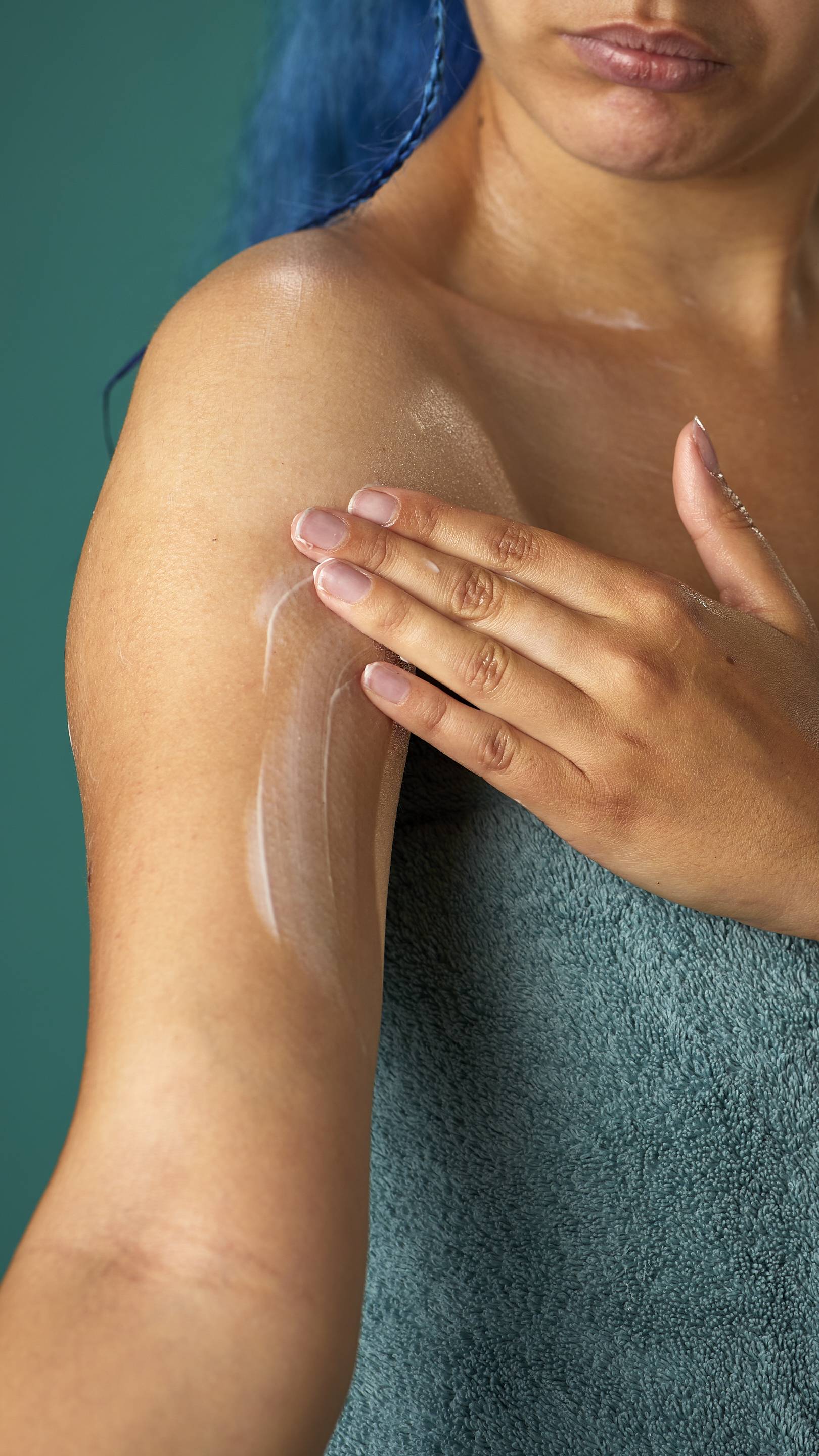 The image shows a close-up of the model, wrapped in a sage-green towel on a teal background. They are gently applying the Coconut body butter to their upper arm.