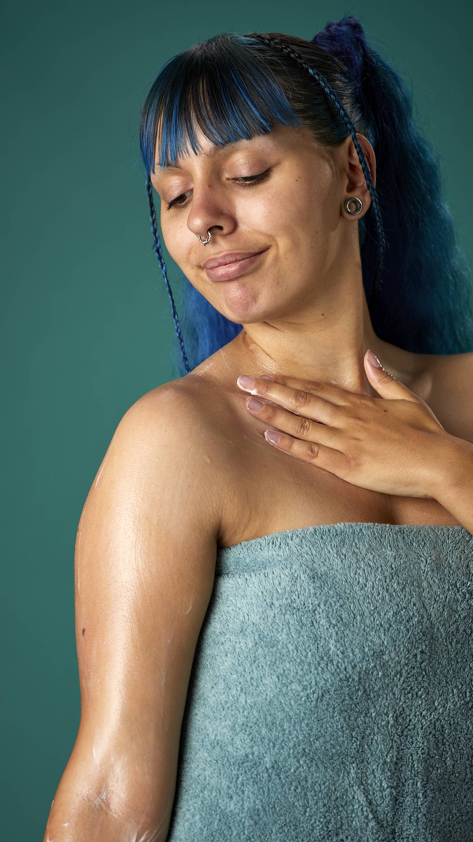 The image shows the model wrapped in a sage-green towel on a teal background. They have blue hair with a full fringe and are gently applying the Coconut body butter to their upper arm.