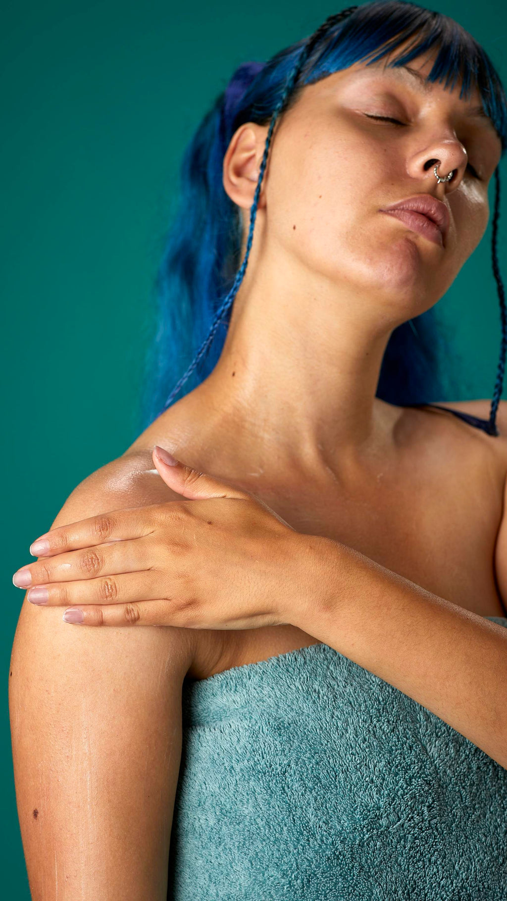 The image shows the model wrapped in a sage-green towel on a teal background. They have blue hair with a full fringe and are gently applying the Coconut body butter to their upper arm.