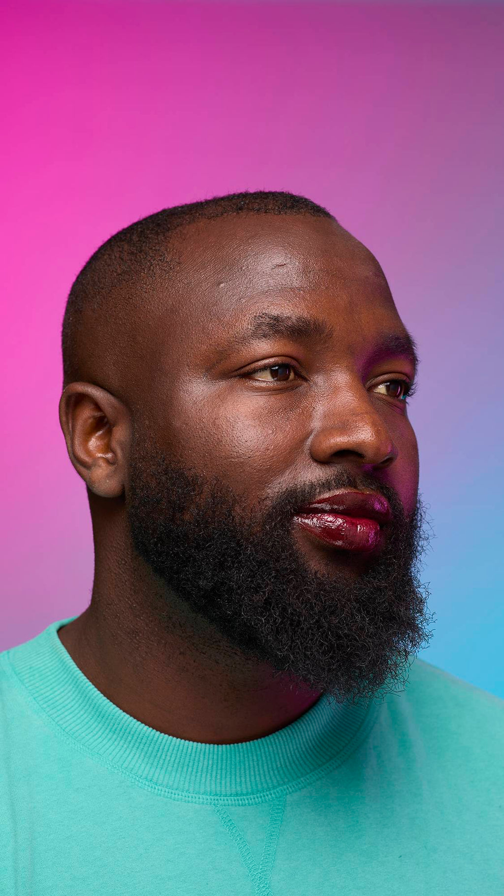 The model is on a pastel pink, blue and purple hue background in a vivid teal jumper. They are glancing away with the focal point being the juicy coating of grounding, earth-toned tint on their lips created by the Cola lip jelly.