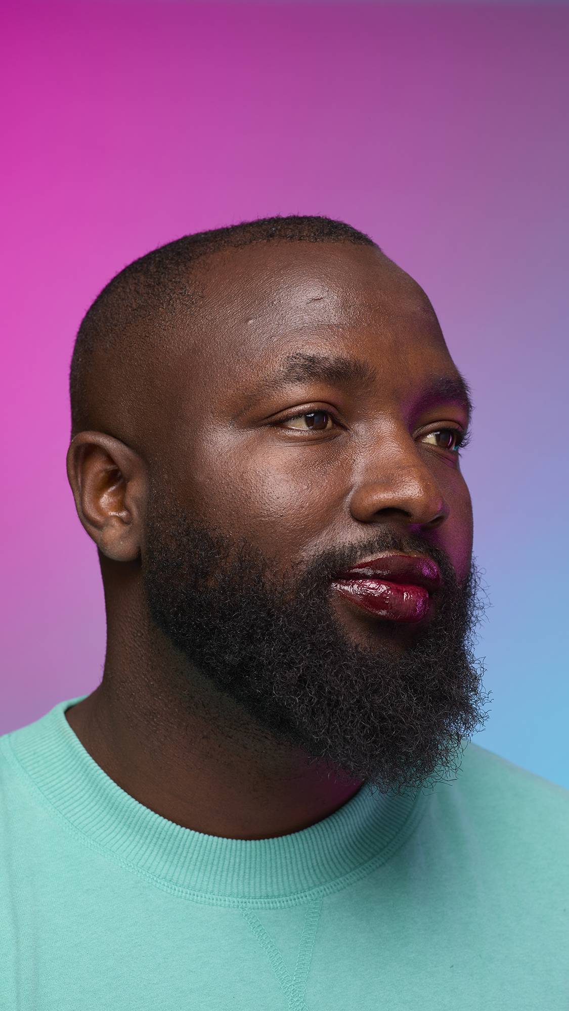 The model is on a pastel pink, blue and purple hue background in a vivid teal jumper. They are glancing away with the focal point being the juicy coating of grounding, earth-toned tint on their lips created by the Cola lip jelly.