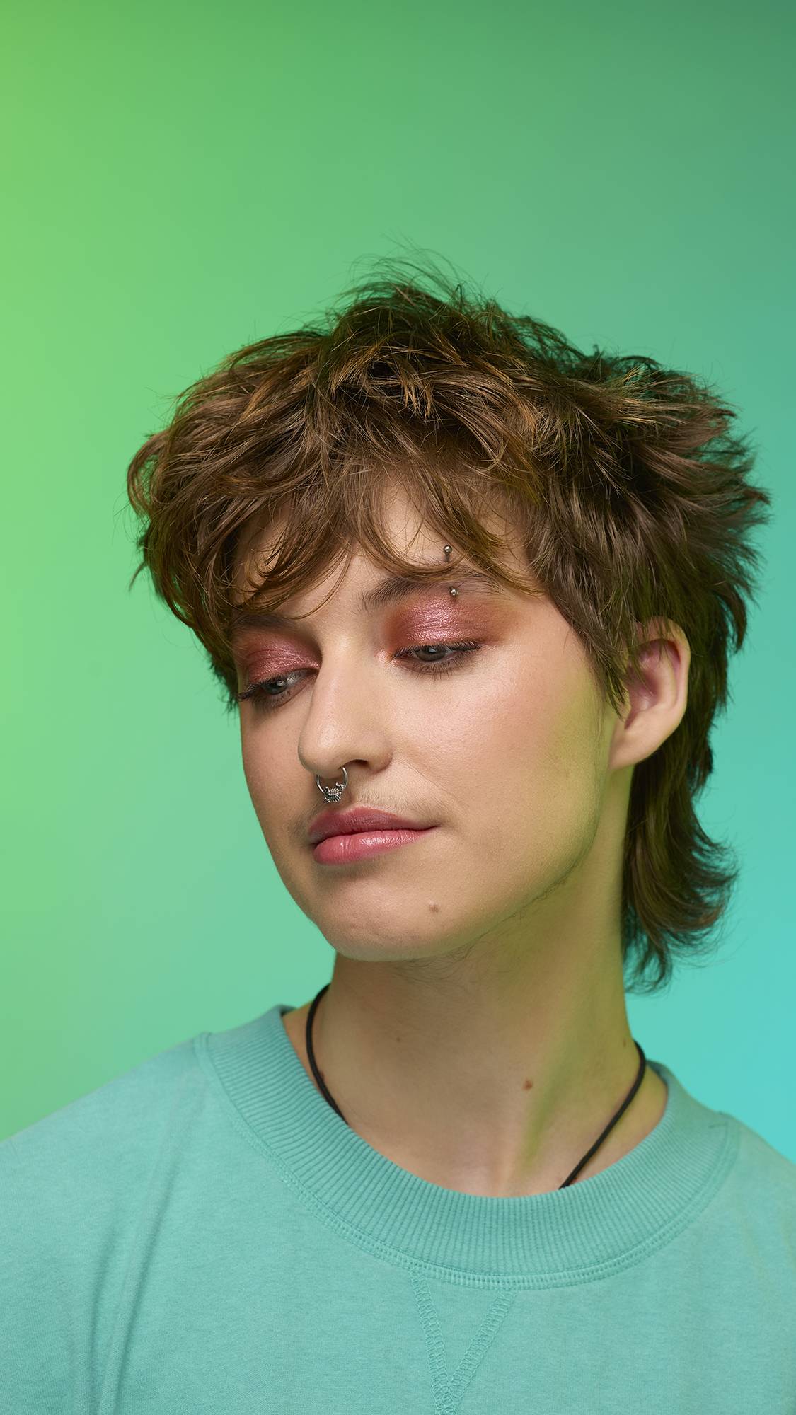 The image shows a close-up of the model's face on a pale blue-to-green gradient background. The model's make-up is done with the focal point being a shimmering, metallic-bronze eyeshadow made by the Comet Glitter Crumble.