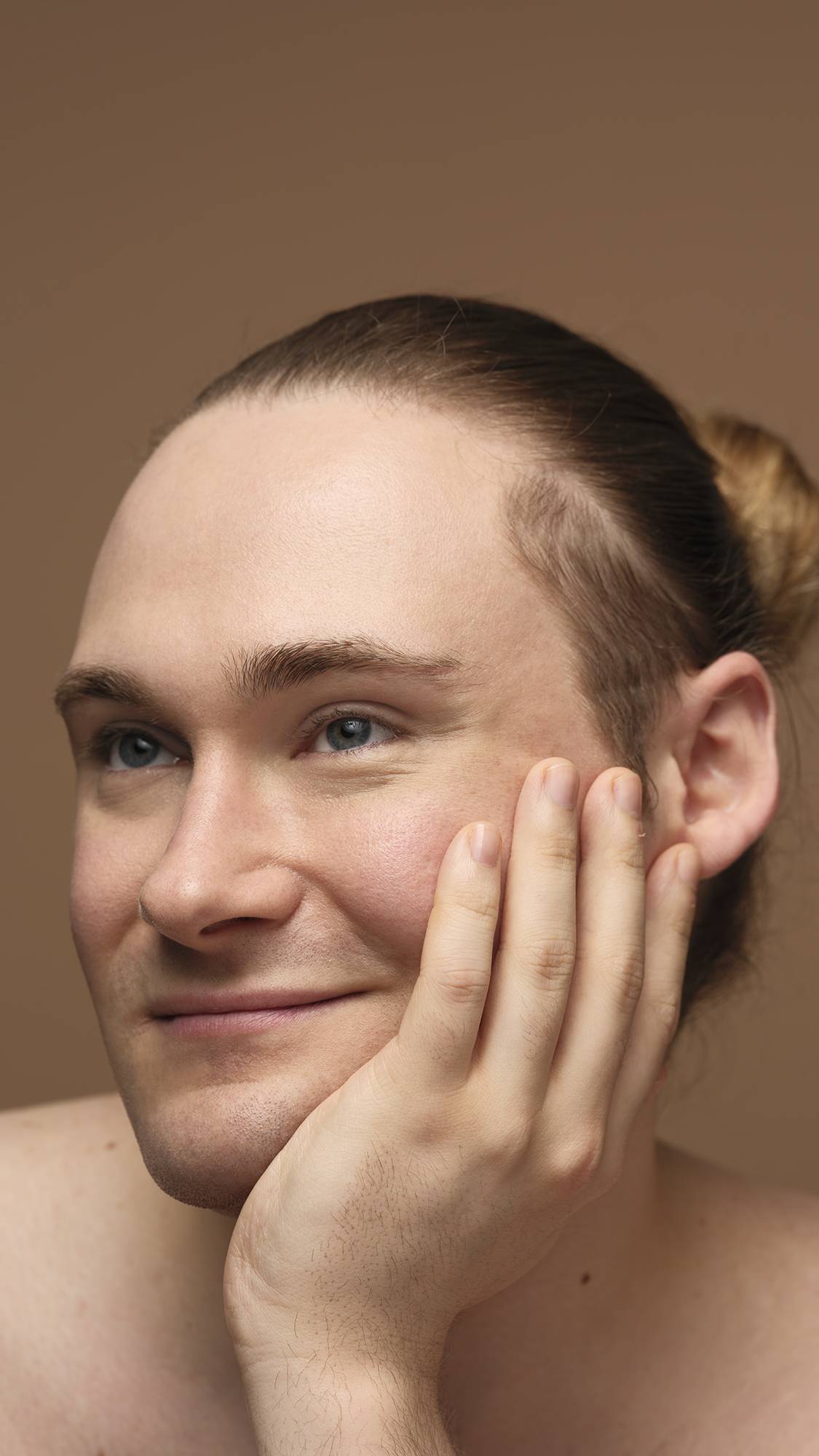 The model is on a warm, earthy-brown background with their head rested upon their hand as they are smiling with glowing, freshly moisturised skin.
