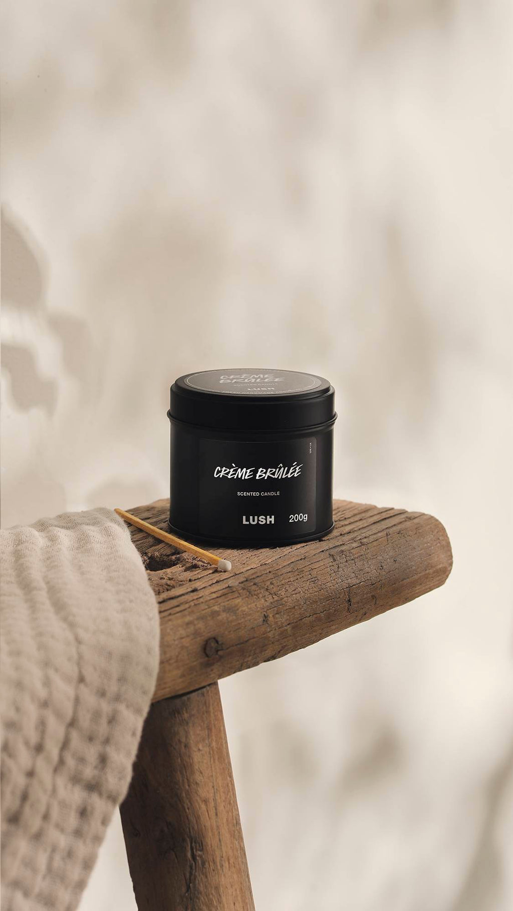 The image shows a blurred, cream background. In the foreground, the Creme Brûlée black tin candle sits on a rustic wooden stool with an unused match beside it and a cream cloth.