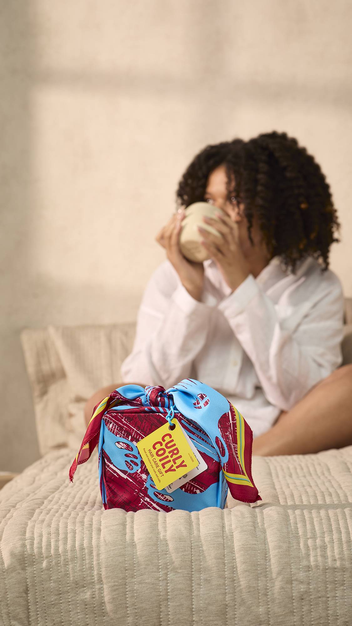 The image shows a light, cream bedroom with the model drinking from a mug in the blurred background as the foreground focuses on the neatly wrapped Curly Coily gift on the end of the bed.