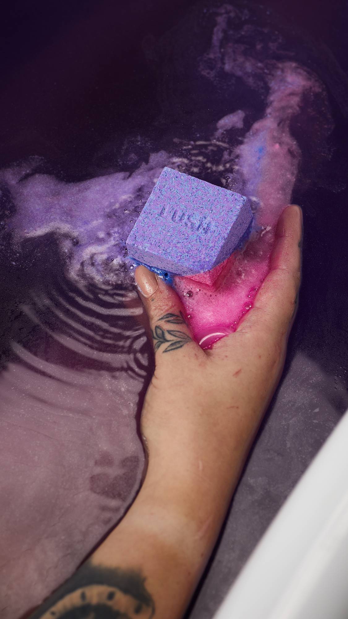 A close-up image of the model's hand gently placing the Lush Deep Sleep Epsom salt cube bath bomb into the water as it begins to give off pink and purple foam.