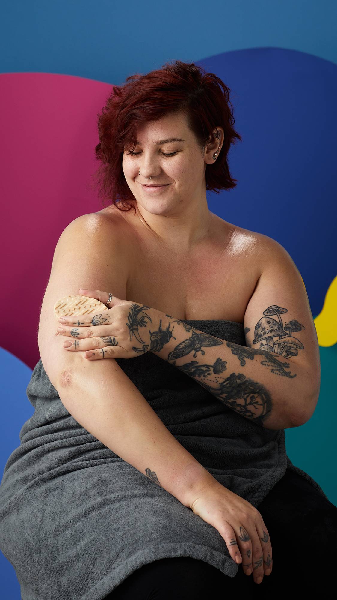 A smiling person with red hair and tattooed arms, wrapped in a grey towel, glides a textured, oval beige Lush massage bar over their arm. They are set against an abstract background of bold, multicoloured shapes.