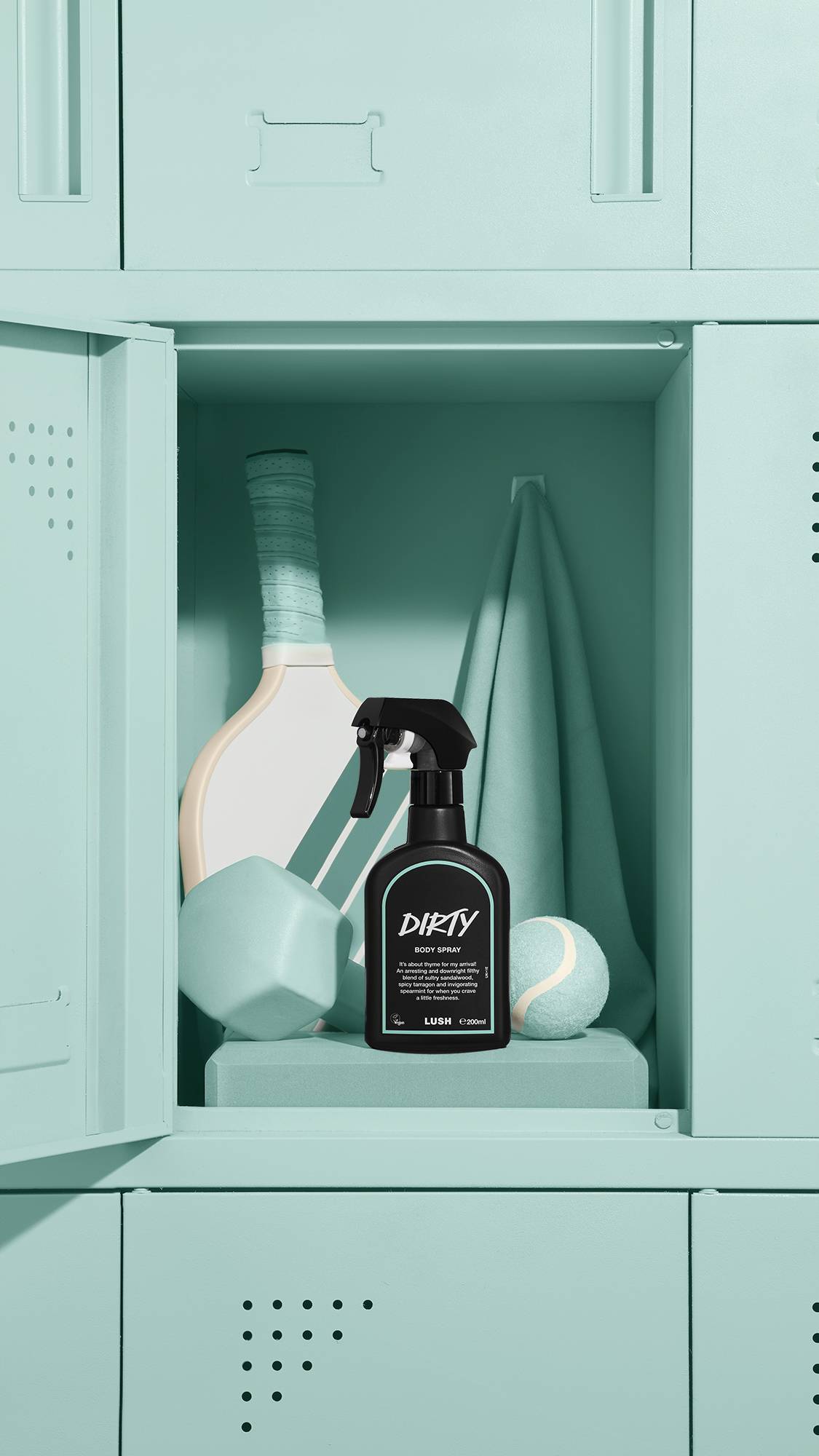 A black bottle of Lush 'Dirty' body spray sits inside an open mint-green locker. Behind the spray are a matching green paddle, towel, tennis ball, and geometric foam blocks, creating a cohesive, sporty aesthetic.