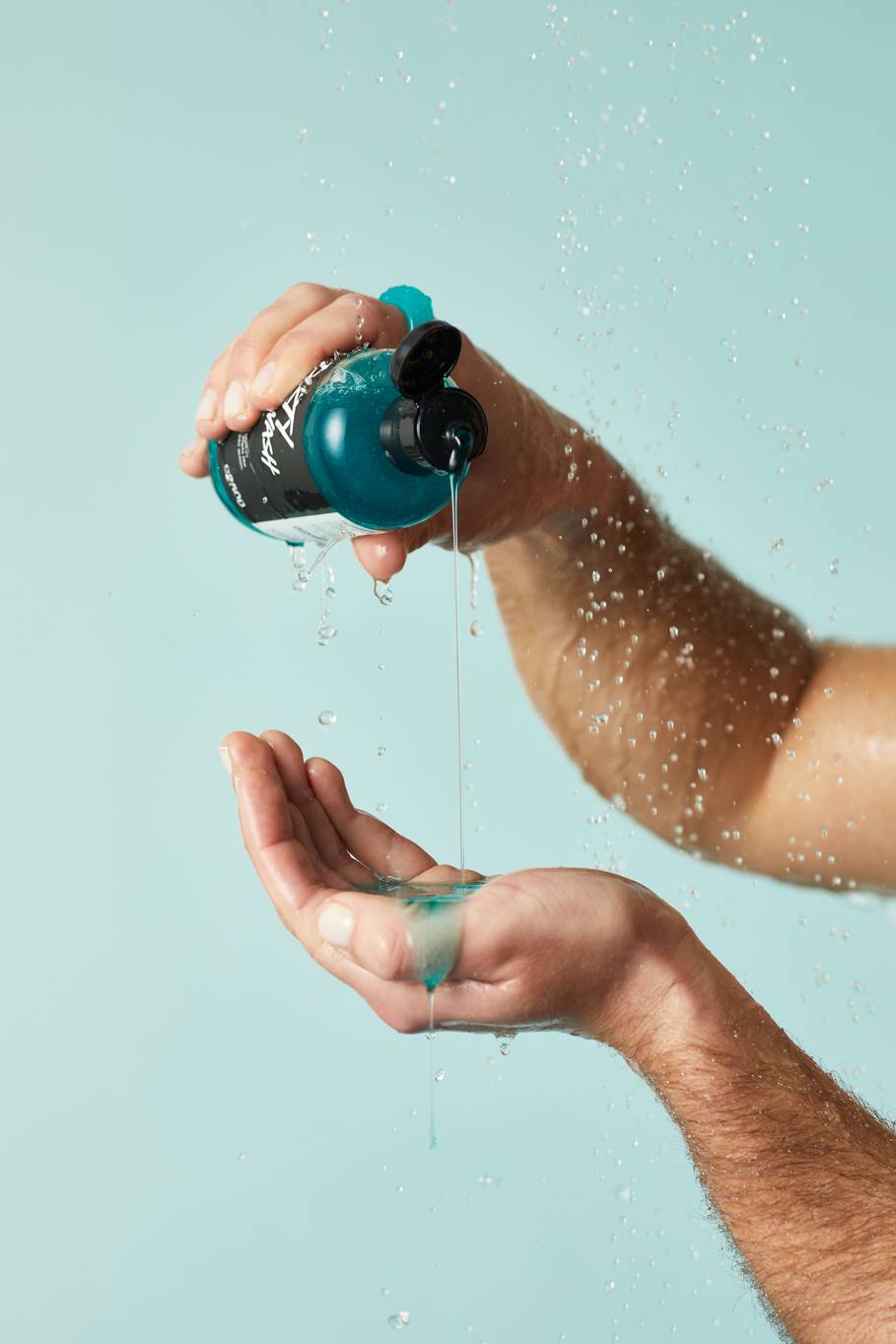 A close-up of the model's forearms on a pastel blue background as they gently squeeze the cool-blue Dirty Springwash shower gel into the palm of their hand under running shower water.