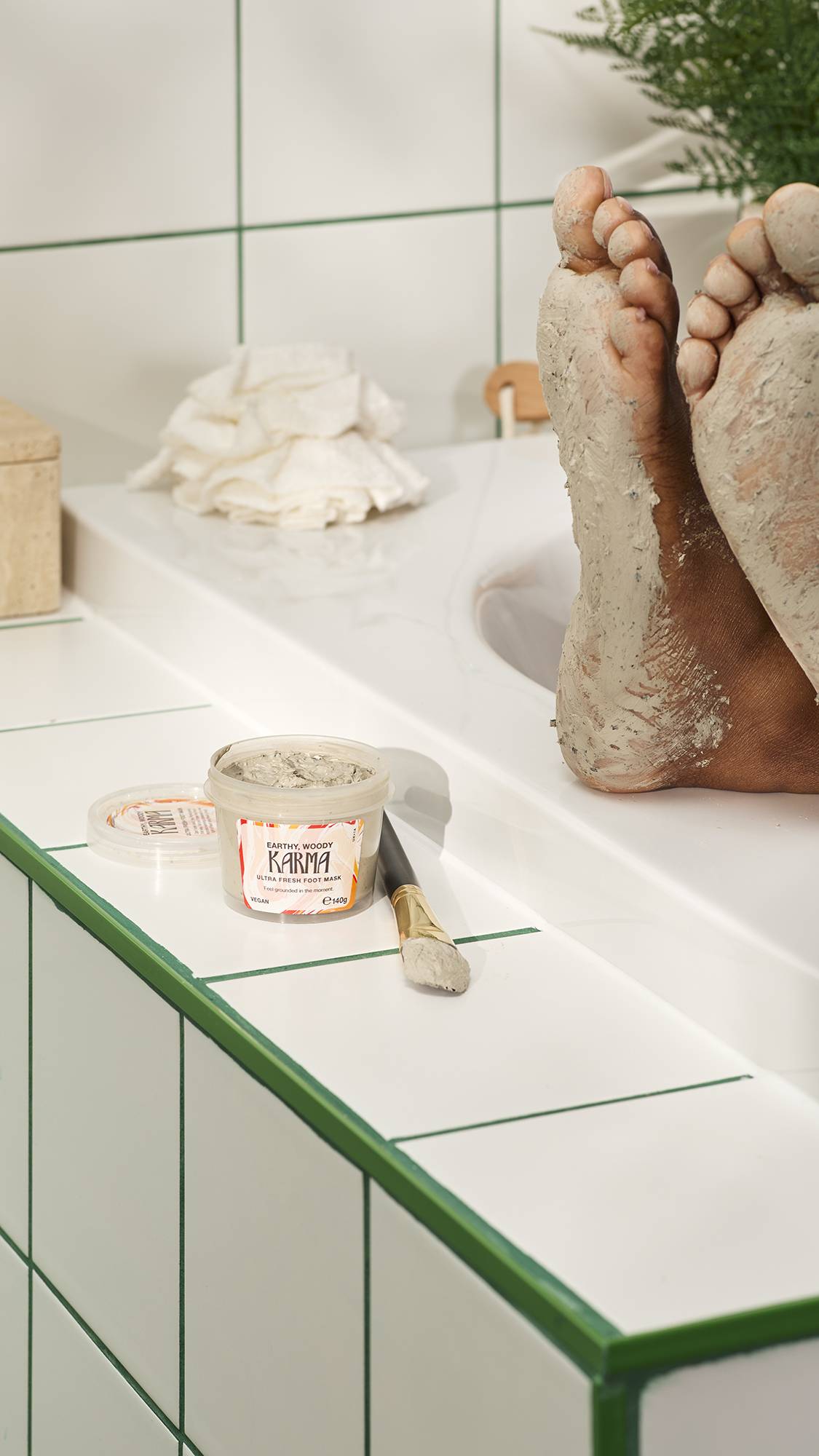 An open pot of Lush "Earthy, Woody Karma" foot mask and a brush rest on a white tiled surface with green grout. In the background, the soles of two feet are coated in the thick, sage-green mask while resting on the edge of a bathtub.