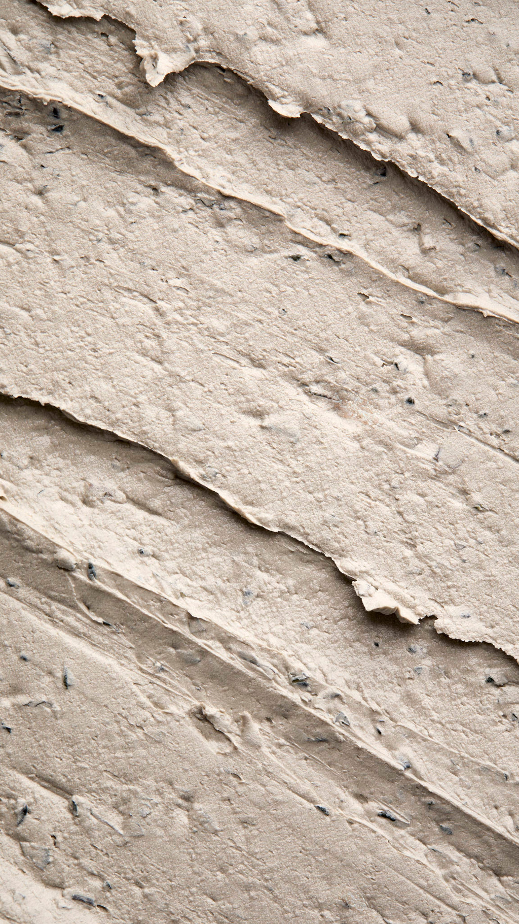 A close-up, textured shot of a thick, "Earthy, Woody Karma" foot mask spread in diagonal ridges. The product has a dense, matte consistency with visible dark exfoliating flecks and small botanical particles throughout.