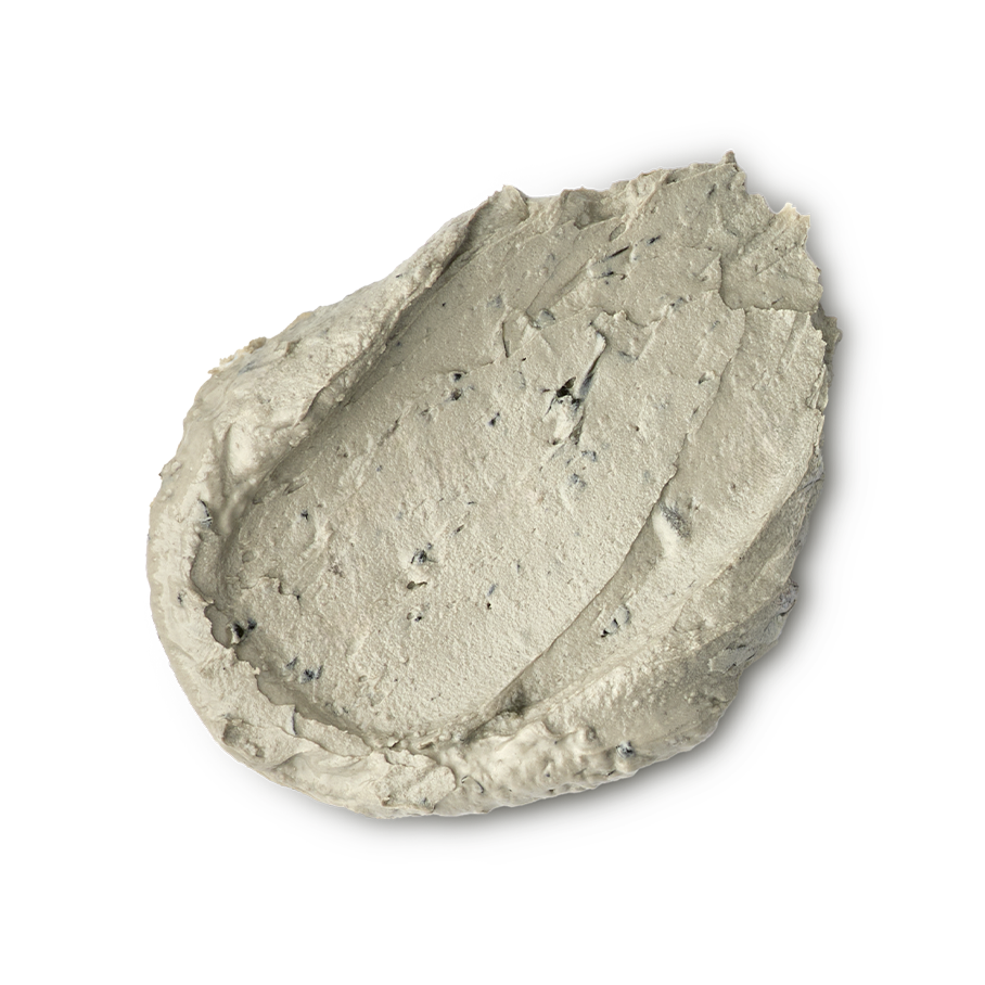 Earthy, Woody, Karma. A thick, textured dollop of pale beige foot mask. The product has a dense, matte consistency with visible dark exfoliating flecks and small lumps, showing a swirled and slightly peaked surface.