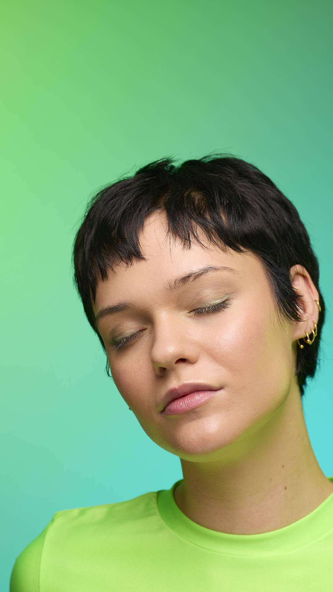 The image shows a close-up of the model's face on a pale blue-to-green gradient background. The model's make-up is done with the focal point being a subtle, gunmetal-brown, shimmering eyeshadow made by the Eclipse Glitter Crumble.