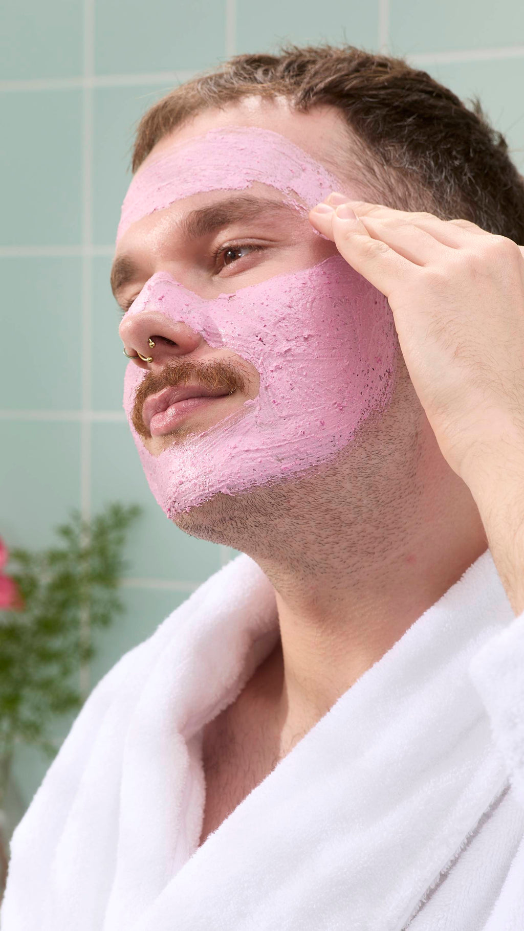 A close-up of a model with a moustache and nose piercings smiling as they apply a thick, grainy pink "Family Face Time" face mask to their forehead. They are wearing a white fluffy dressing gown against a background of pale green tiles.