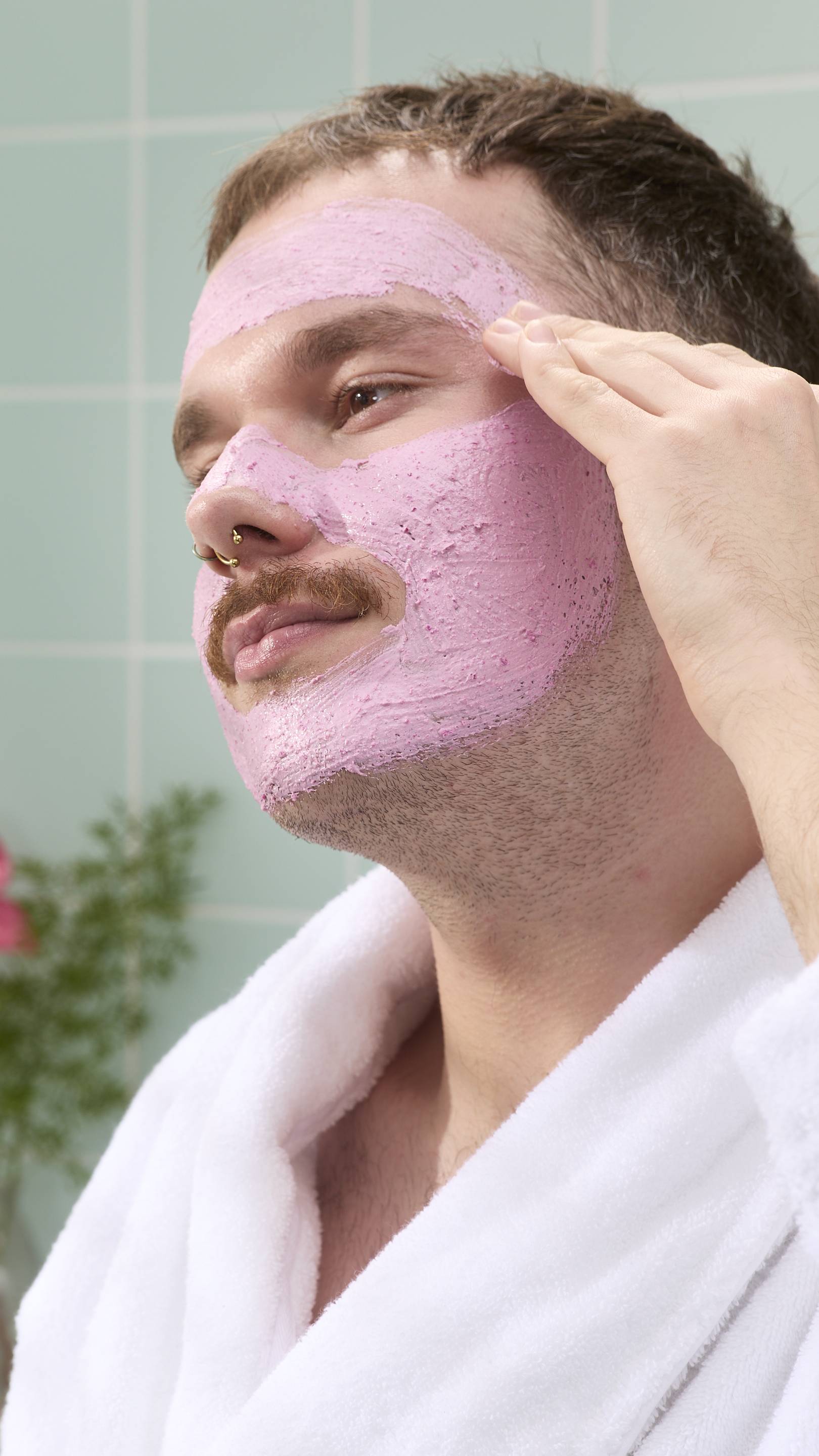 A close-up of a model with a moustache and nose piercings smiling as they apply a thick, grainy pink "Family Face Time" face mask to their forehead. They are wearing a white fluffy dressing gown against a background of pale green tiles.