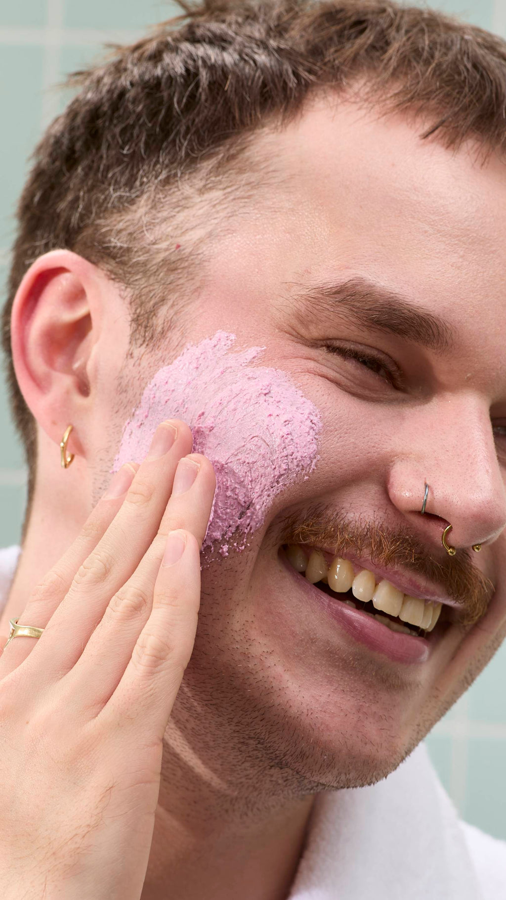 A model with a moustache and nose piercings smiles while applying a thick, grainy pink "Family Face Time" face mask to their cheek. They are wearing a white dressing gown against a background of pale green tiles.