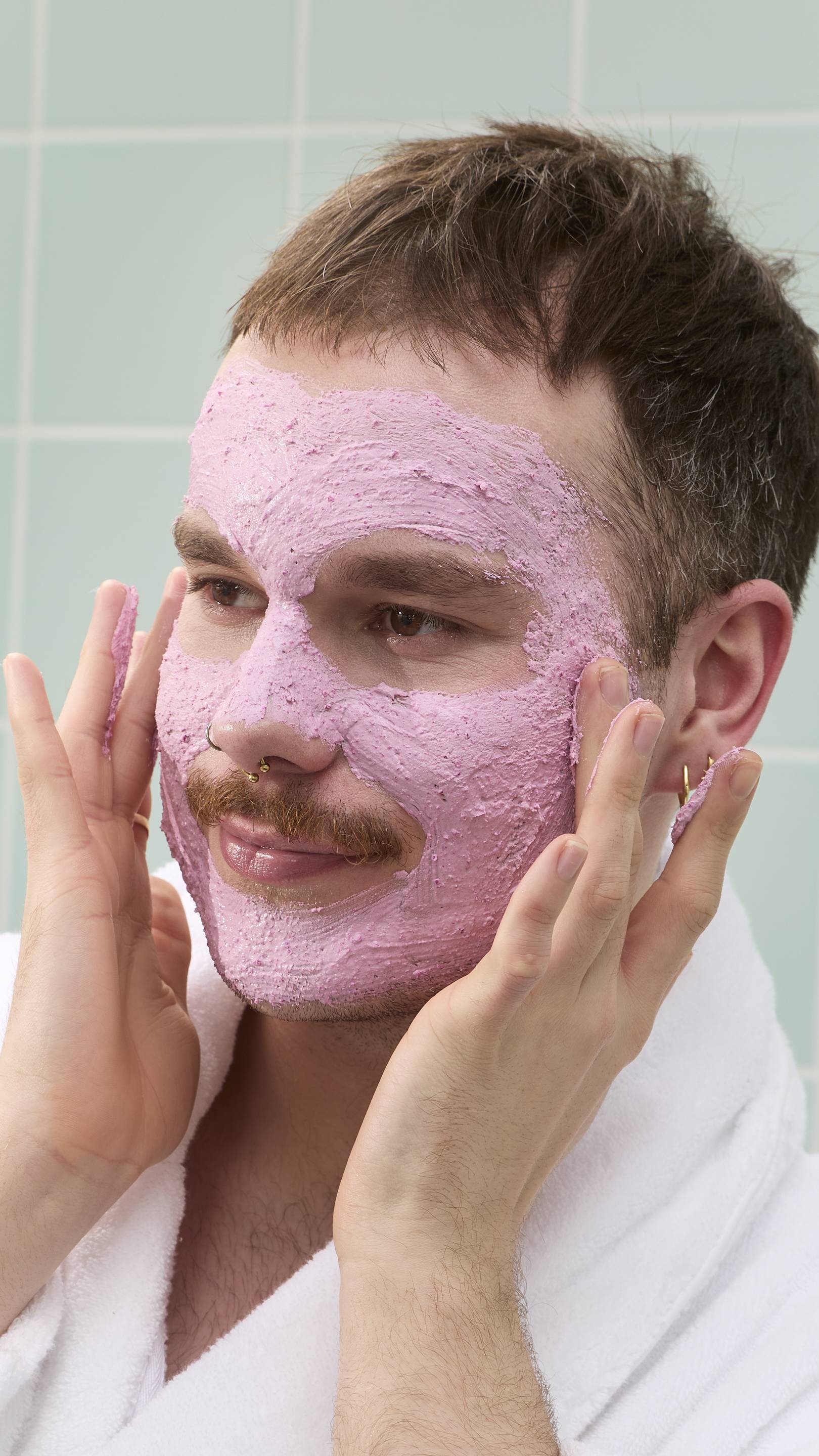 A model with a moustache and nose piercings smiles while looking to the side. Most of their face is covered in a thick, pink "Family Face Time" face mask. They wear a white dressing gown against a background of pale green bathroom tiles.