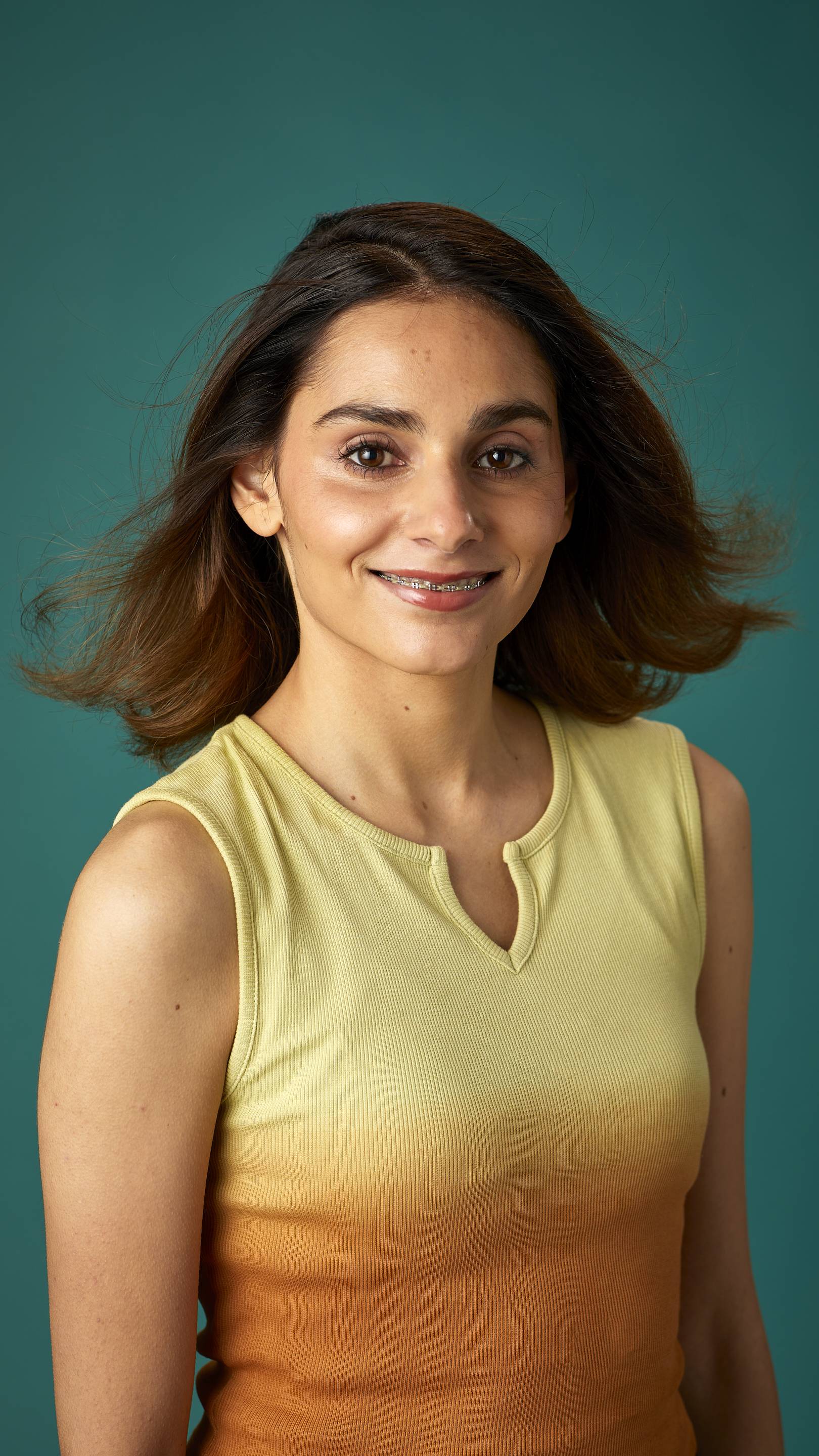 The model is on a teal background with a contrasting, yellow and orange gradient tank top. They have thick, shoulder-length hair which looks clean and healthy and is being fanned with air.