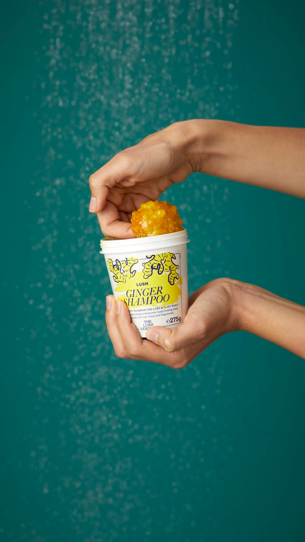 The image shows the model's arms stretched into the centre holding the white, LUSH Version-themed packed pot of the Ginger shampoo. One of their hands is scooping the product out. There is a teal background.
