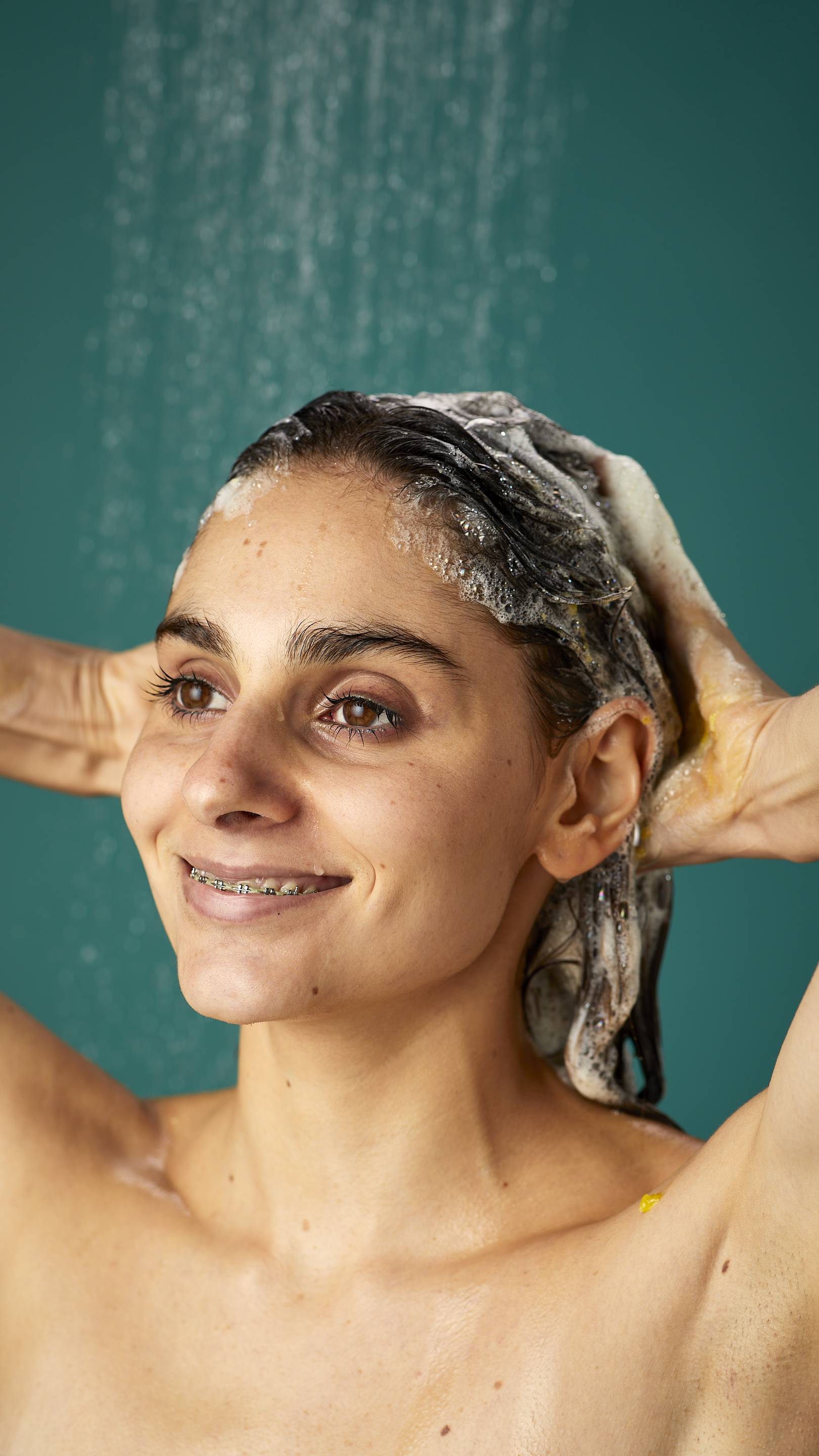 The model is standing under running shower water on a teal background. Their dark, long hair is wet and lathered in suds as they lather the Ginger shampoo into their roots.