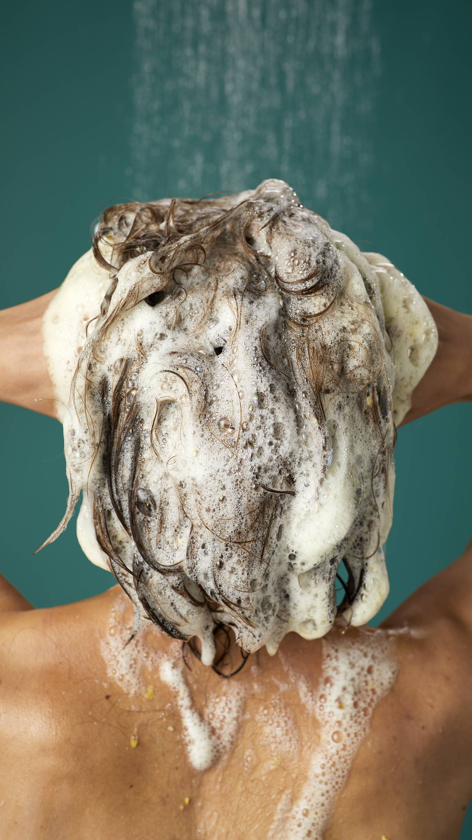 The image shows the rear view of the model's dark, long hair which is wet and lathered in suds as they lather the Ginger shampoo into their roots. There is a teal background with running shower water.