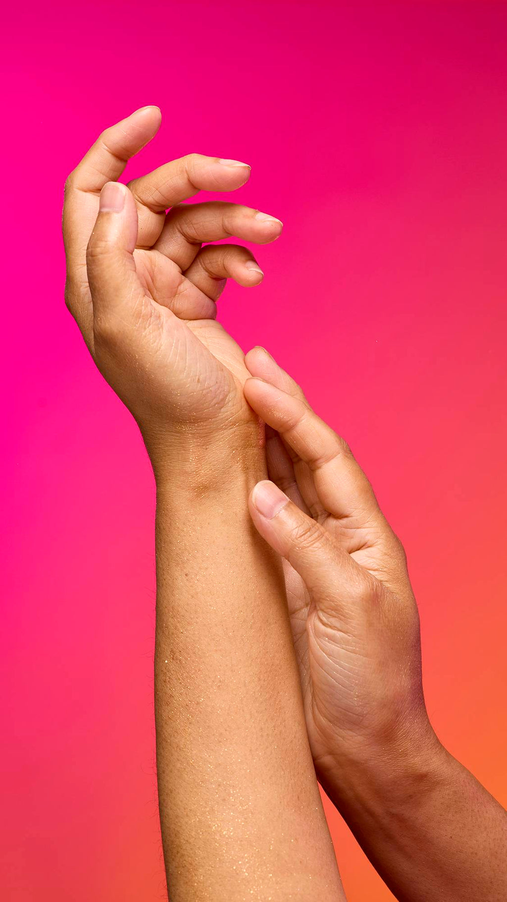 The image shows a model's forearm extended from the bottom as the skin appears moisturised with a subtle, delicate shimmer over the skin after having used the Golden Egg Glitter Mist spray product on a pink-to-orange gradient background.
