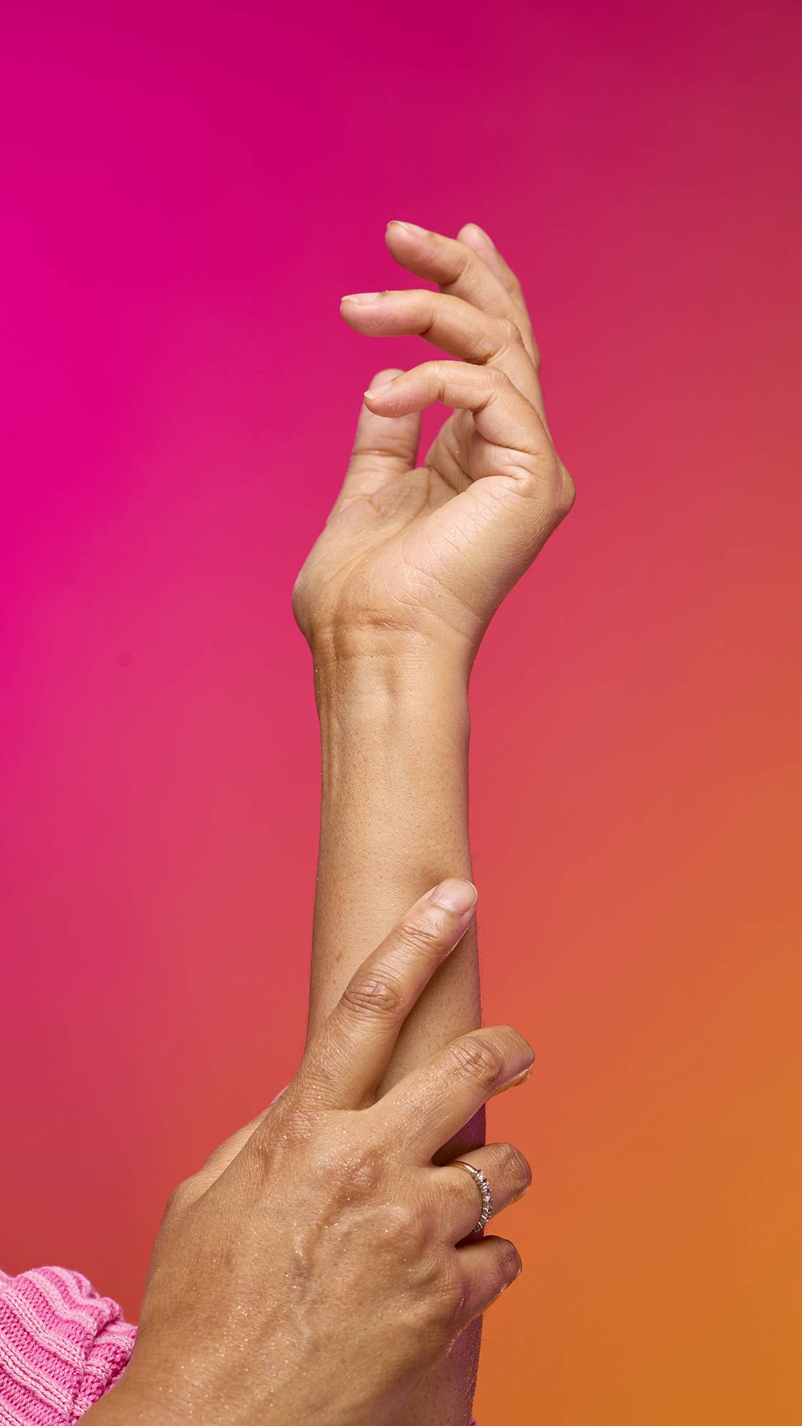 The image shows a model's forearm extended from the bottom as the skin appears moisturised with a subtle, delicate shimmer over the skin after having used the Golden Egg Glitter Mist spray product on a pink-to-orange gradient background.