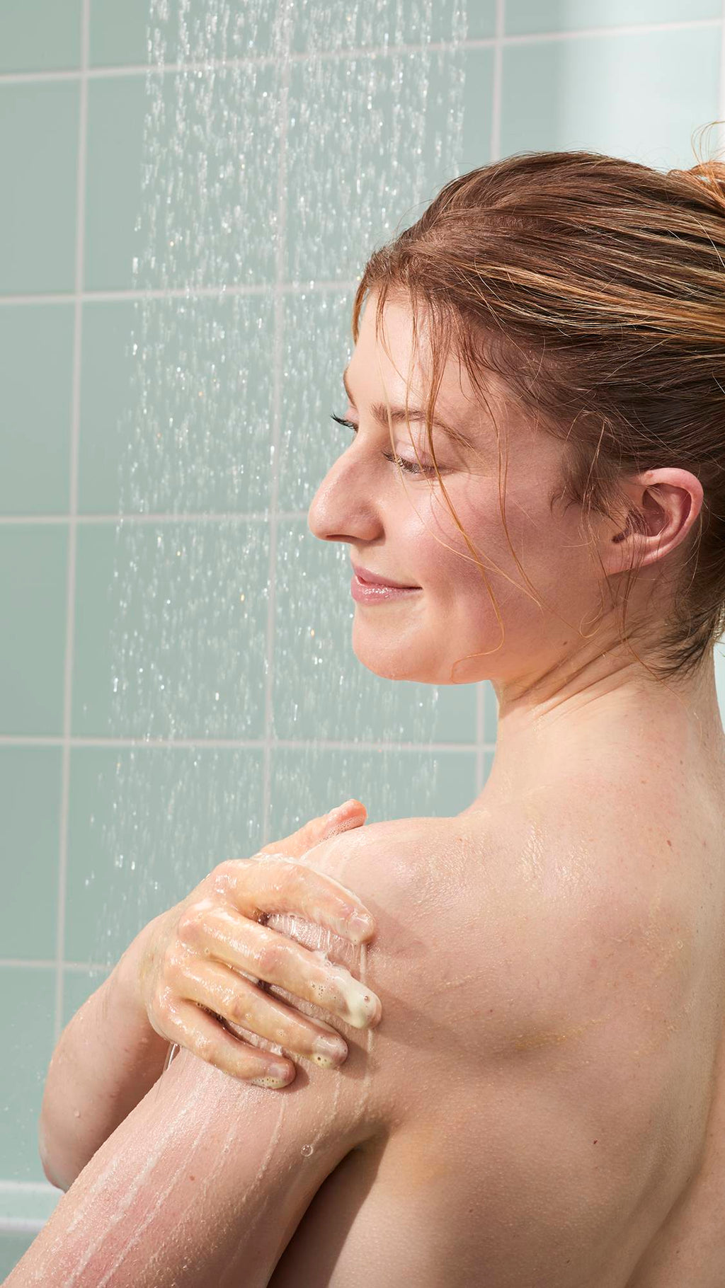 A woman with reddish-brown hair smiles in the shower while lathering a shimmering gold gel onto her shoulder. The background features light mint green tiles and the soft, vertical spray of falling water.