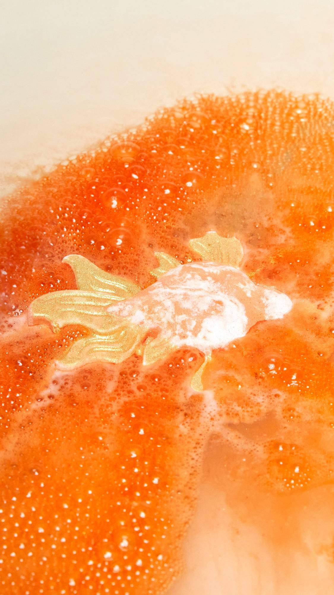 The Goldfish bath bomb is slowly dissolving creating a shimmering sea of deep, sunset-orange water and thick, vibrant foam.