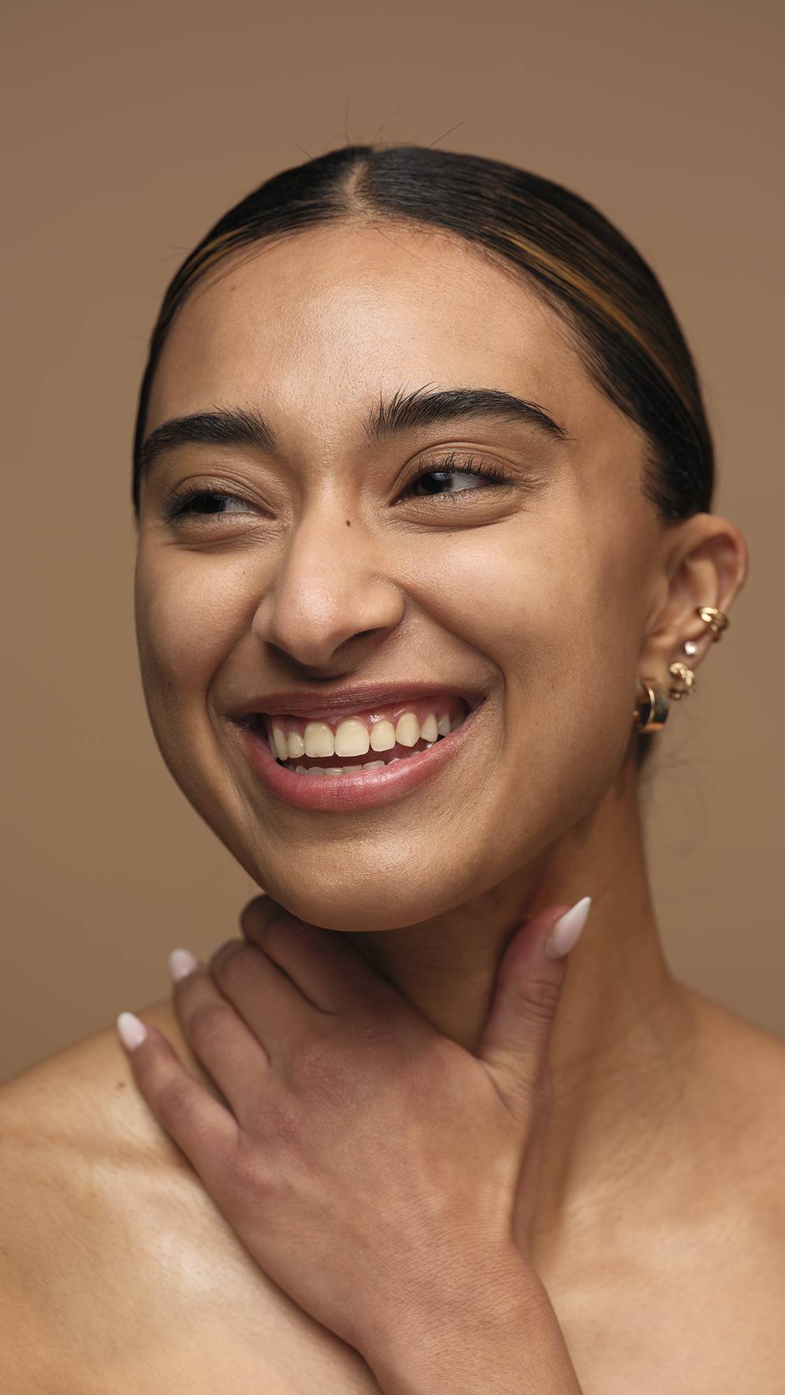 The model is on a warm, earthy-brown background with their hand across their chest and neck as they are smiling with glowing, freshly moisturised skin.