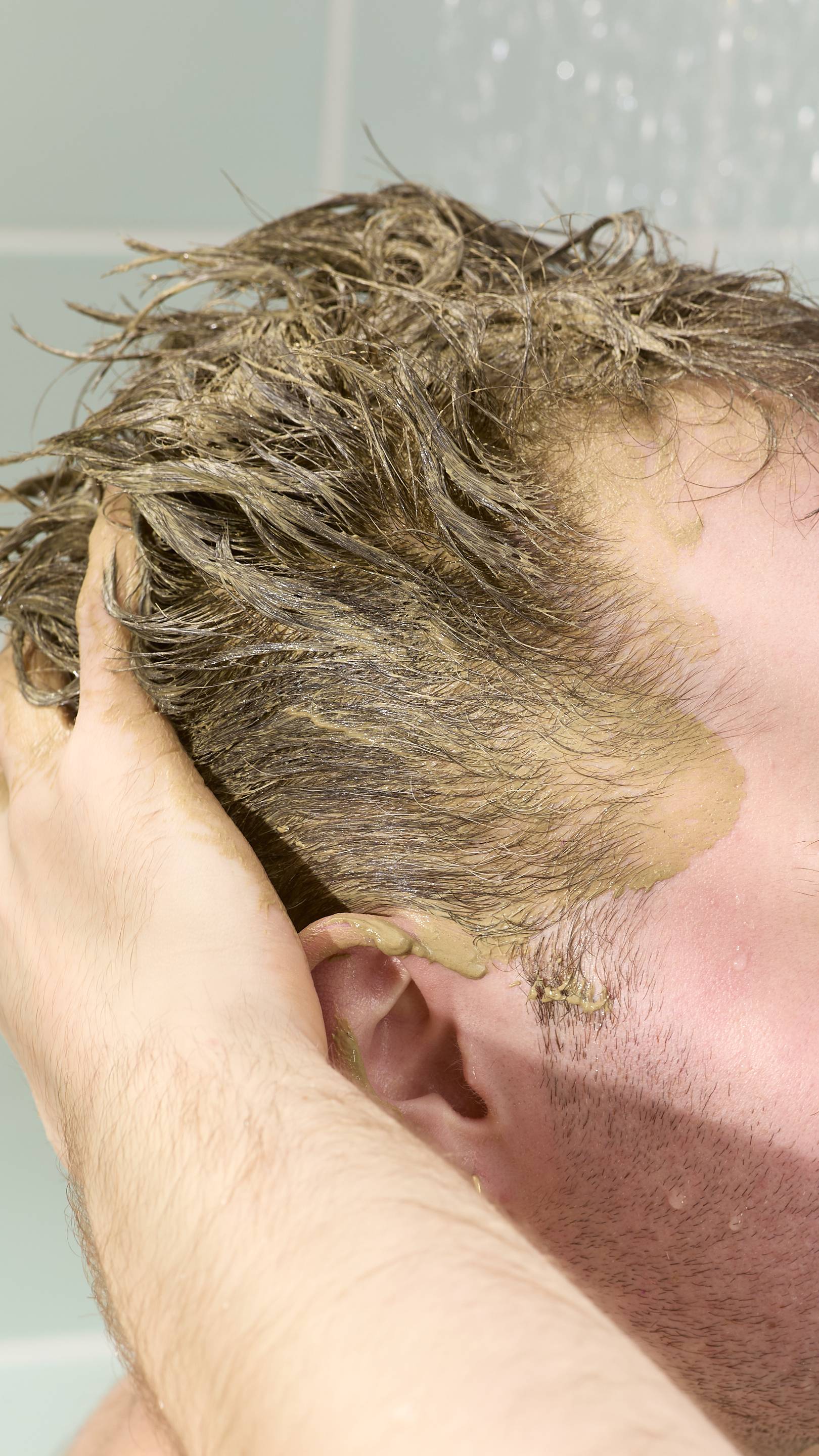 A close-up of a model applying a thick, olive-green "Green Shoots" hair food treatment to their short, dark blonde hair. Their hand is pressed against the side of their head, with the paste visible on their temple, ear, and through the hair strands.