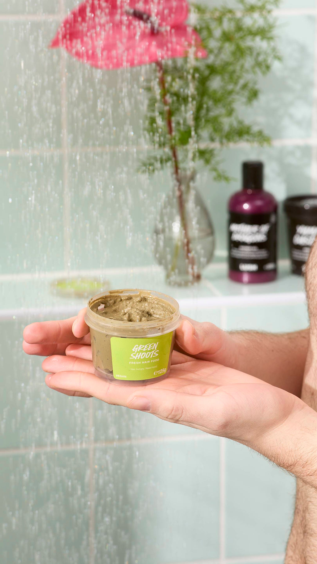 A close-up of a model holding an open pot of "Green Shoots" fresh hair food in their hands. They are standing in a shower with water falling in the foreground. In the background, there is a pink flower and other skincare products on a tiled ledge.