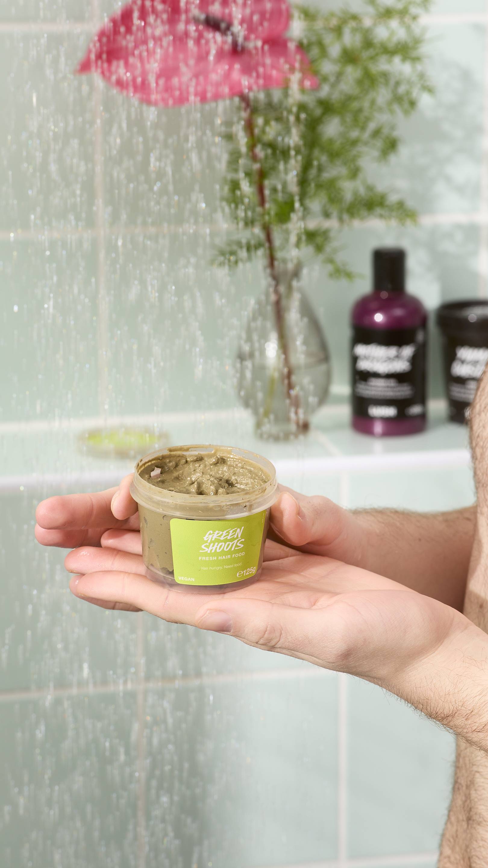 A close-up of a model holding an open pot of "Green Shoots" fresh hair food in their hands. They are standing in a shower with water falling in the foreground. In the background, there is a pink flower and other skincare products on a tiled ledge.