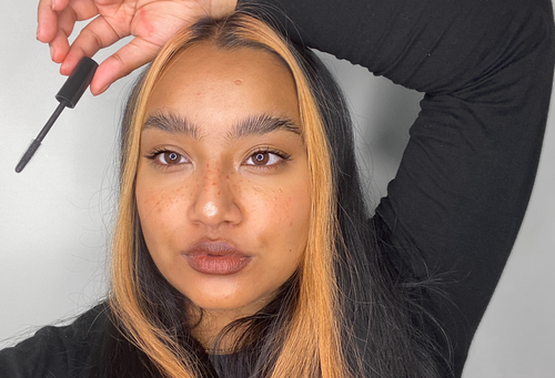 A person wearing mascara pouts and poses with their weapon of choice; a Lush Lashes mascara wand, featuring super fine bristles.