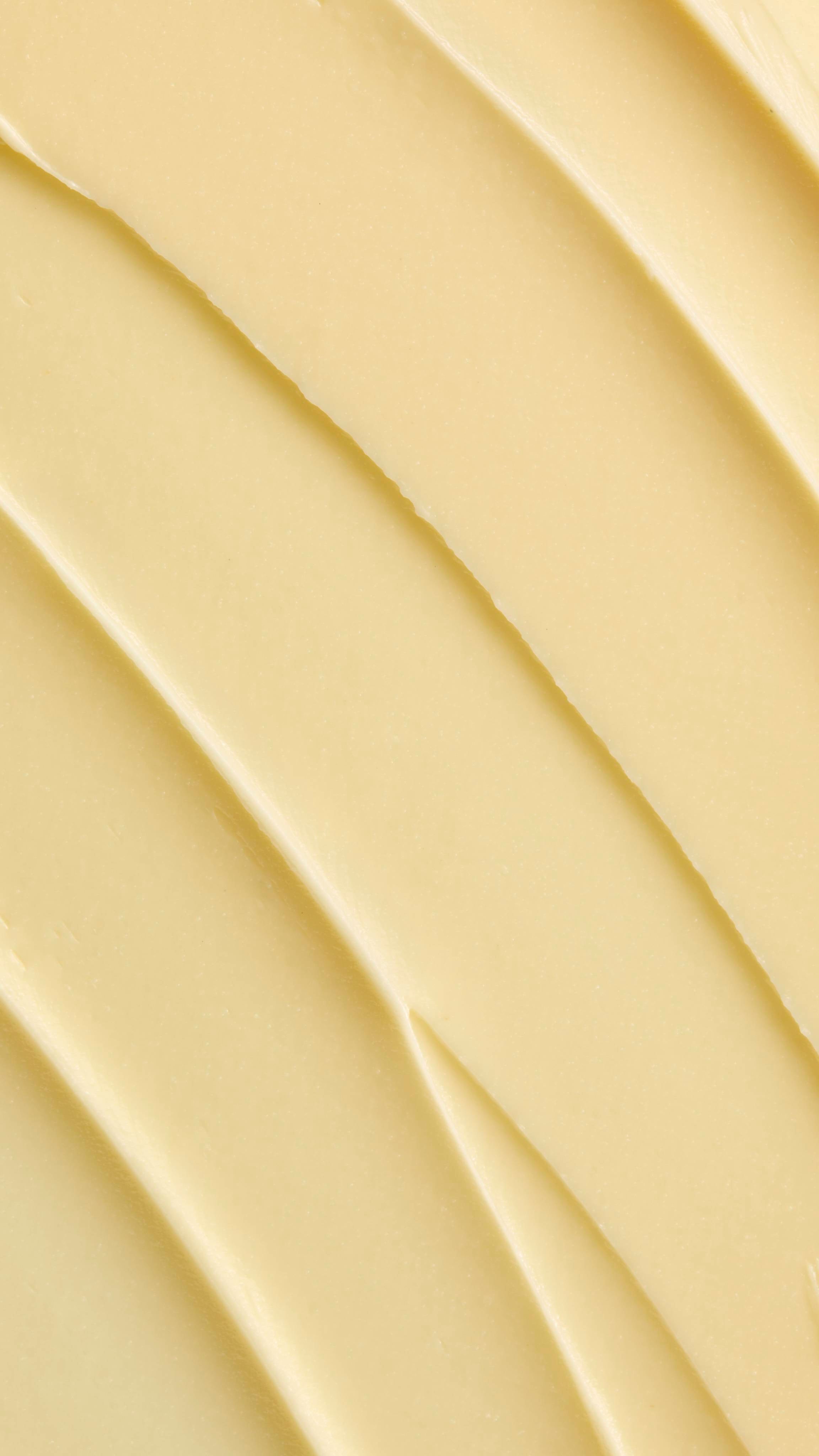 A super close-up image showing diagonal, overlapping smears of the thick, creamy, pale yellow Lush "Honey, Beeswax And Almond Oil" facial cleanser. The smooth product has a matte finish and subtle ridged textures.