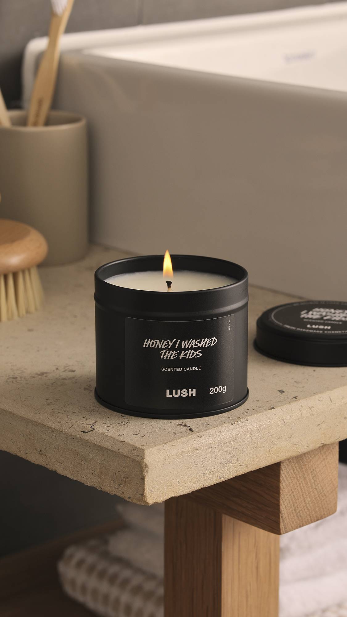 The image shows a blurred background with a white, square bath and bamboo toothbrush. In the foreground, the lit Honey I Washed The Kids black tin candle sits atop a wooden, stone-topped counter.