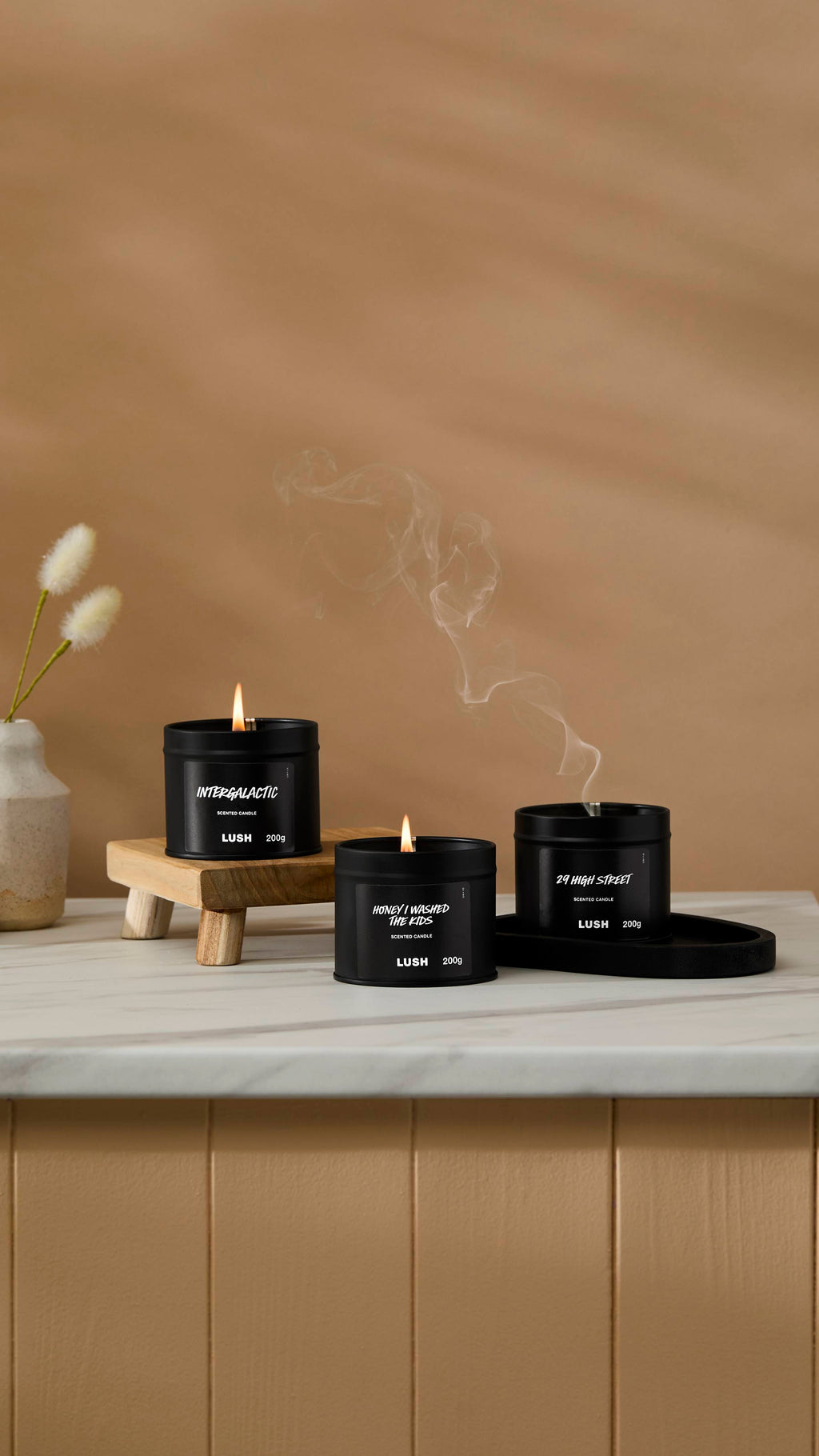 The image shows a warm beige background with a ceramic vase of Hare's Tail Grass. Three back tin candles are sitting with their wicks lit on a marble-topped counter with one having just been blown out with smoke rising,