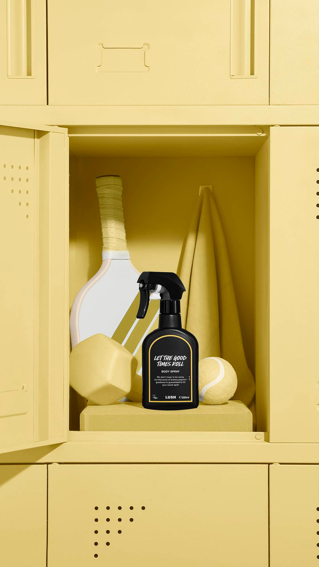A black Lush body spray bottle labelled 'Let The Good Times Roll' sits inside an open yellow locker. The locker also contains a sports racket, a yellow tennis ball, a yellow geometric shape, and a hanging towel.