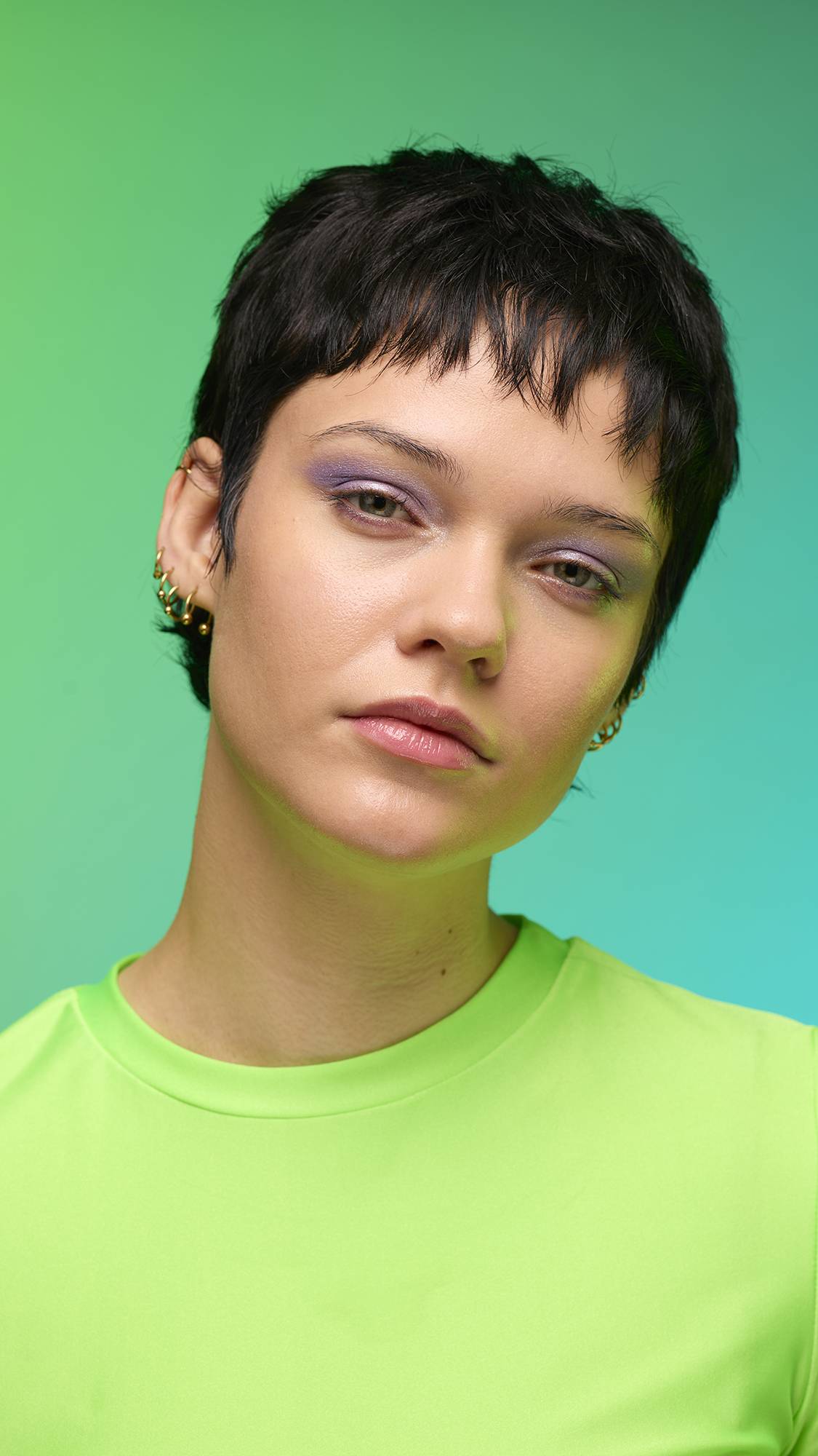 The image shows a close-up of the model's face on a pale blue-to-green gradient background. The model's make-up is done with the focal point being a delicate, purple, shimmering eyeshadow made by the Little Sparkler Glitter Crumble.