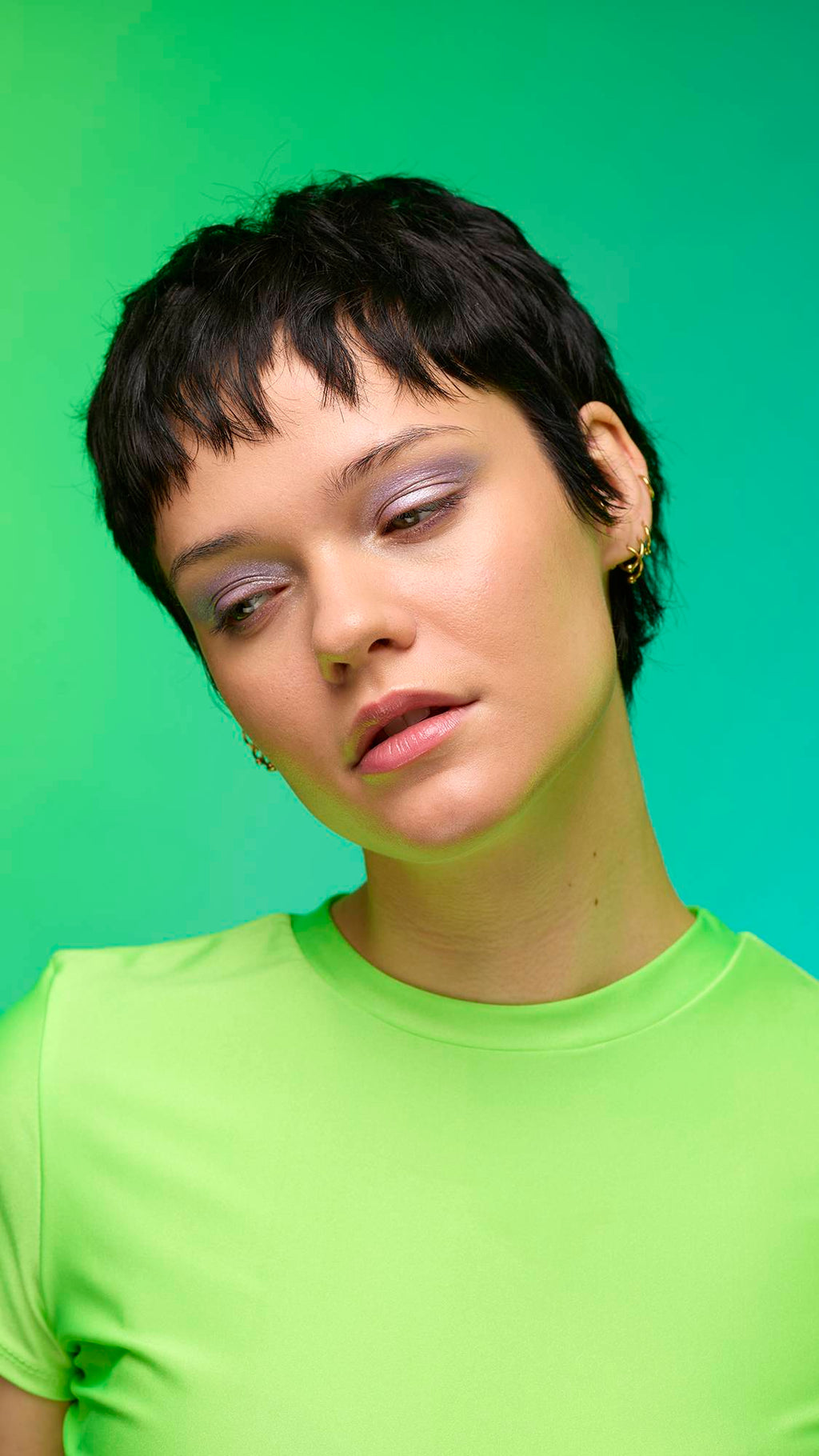 The image shows a close-up of the model's face on a pale blue-to-green gradient background. The model's make-up is done with the focal point being a delicate, purple, shimmering eyeshadow made by the Little Sparkler Glitter Crumble.