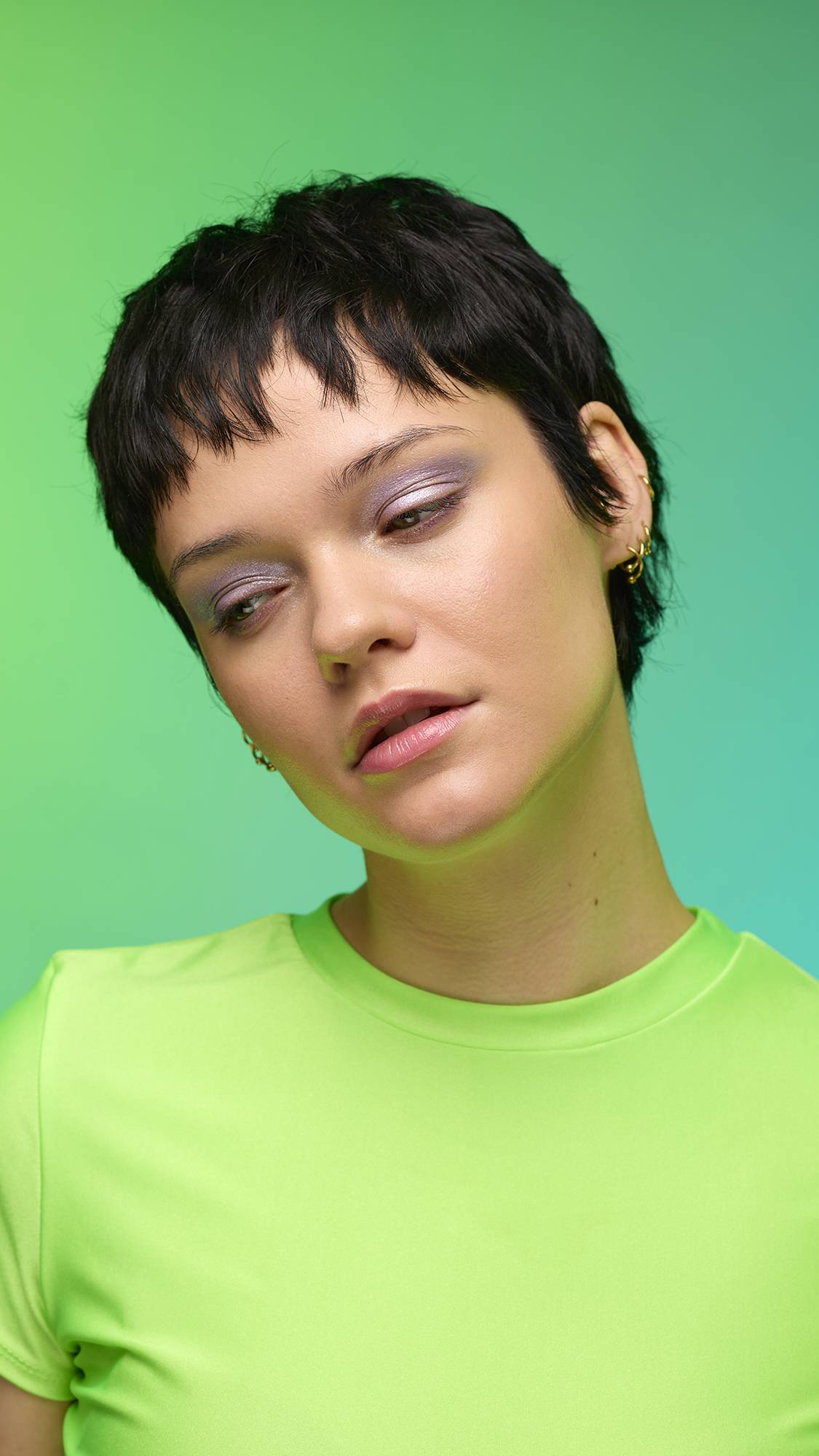 The image shows a close-up of the model's face on a pale blue-to-green gradient background. The model's make-up is done with the focal point being a delicate, purple, shimmering eyeshadow made by the Little Sparkler Glitter Crumble.