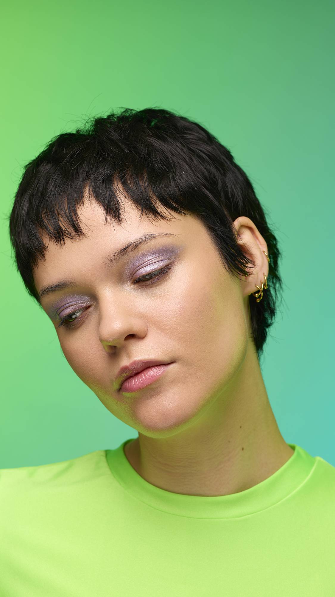 The image shows a close-up of the model's face on a pale blue-to-green gradient background. The model's make-up is done with the focal point being a delicate, purple, shimmering eyeshadow made by the Little Sparkler Glitter Crumble.