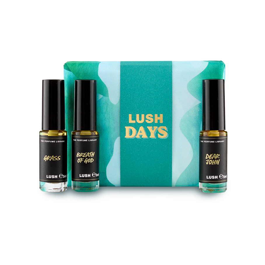The image shows a "LUSH DAYS" perfume gift set. Three cylindrical glass perfume bottles are arranged around a vibrant teal box with a wavy pattern and yellow text. Perfumes include 'Grass', 'Breath of God' and 'Dear John'.