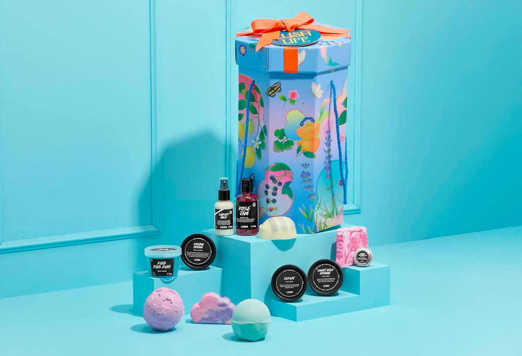 The image shows a tall, hexagonal 'Lush Life' gift box with colourful illustrations, sitting on a blue plinth. A selection of 12 products are arranged on steps around it, against a light blue panelled wall.