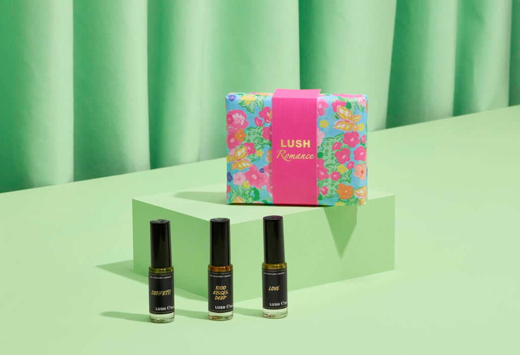 The image shows a "LUSH Romance" perfume gift set: three thin glass bottles on light green steps. A vibrant, floral, light blue box with a bright pink band sits behind them. Matching green fabric forms the background.