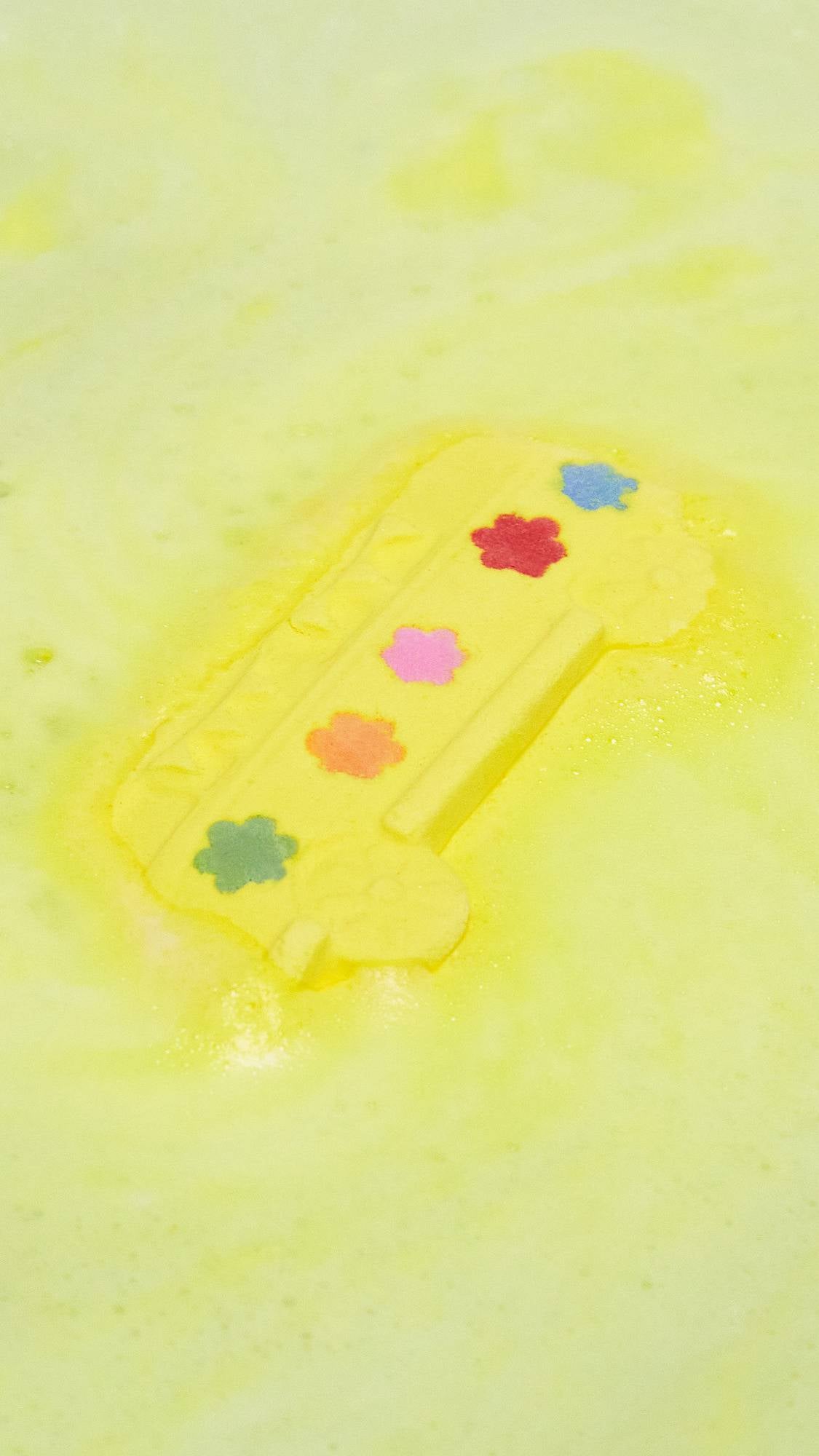 An aerial view image of the Magic Bus, yellow school bus-shaped bath bombs with colourful flower detail on the side as it dissolves in the bath. The product creates a thick layer of velvety, canary-yellow foam across the surface of the water.