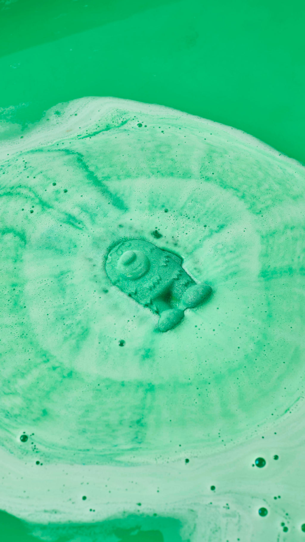 A mint-green, monster-shaped bath bomb dissolves at the centre of a bath, generating concentric ripples of lime-green and pale-green foam. The surrounding water is a vivid, opaque shade of green.