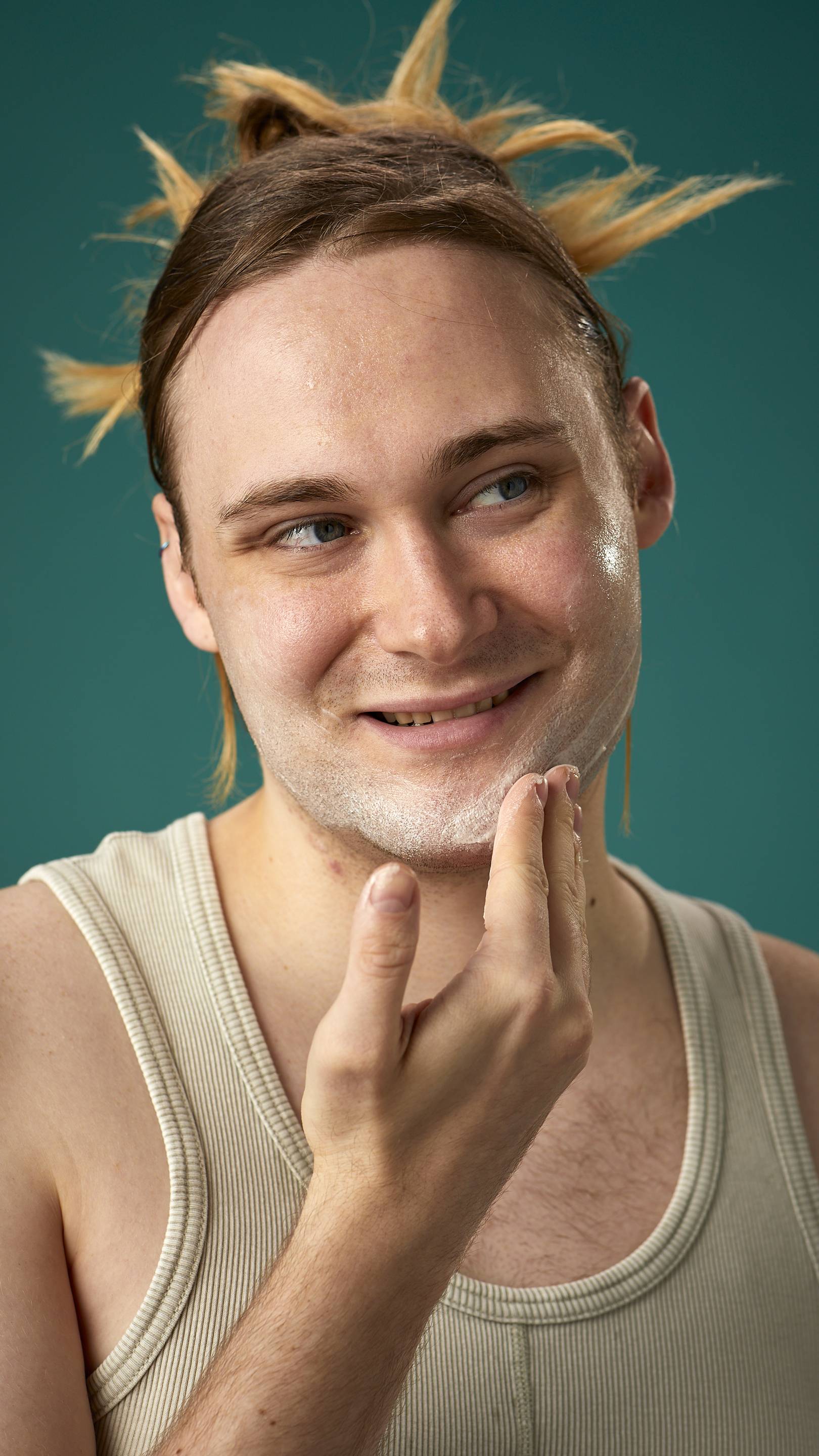 The image shows the model on a teal background with their hair tied back. They have a clean, fresh face as they gently apply the Mostly facial wash over their cheeks.