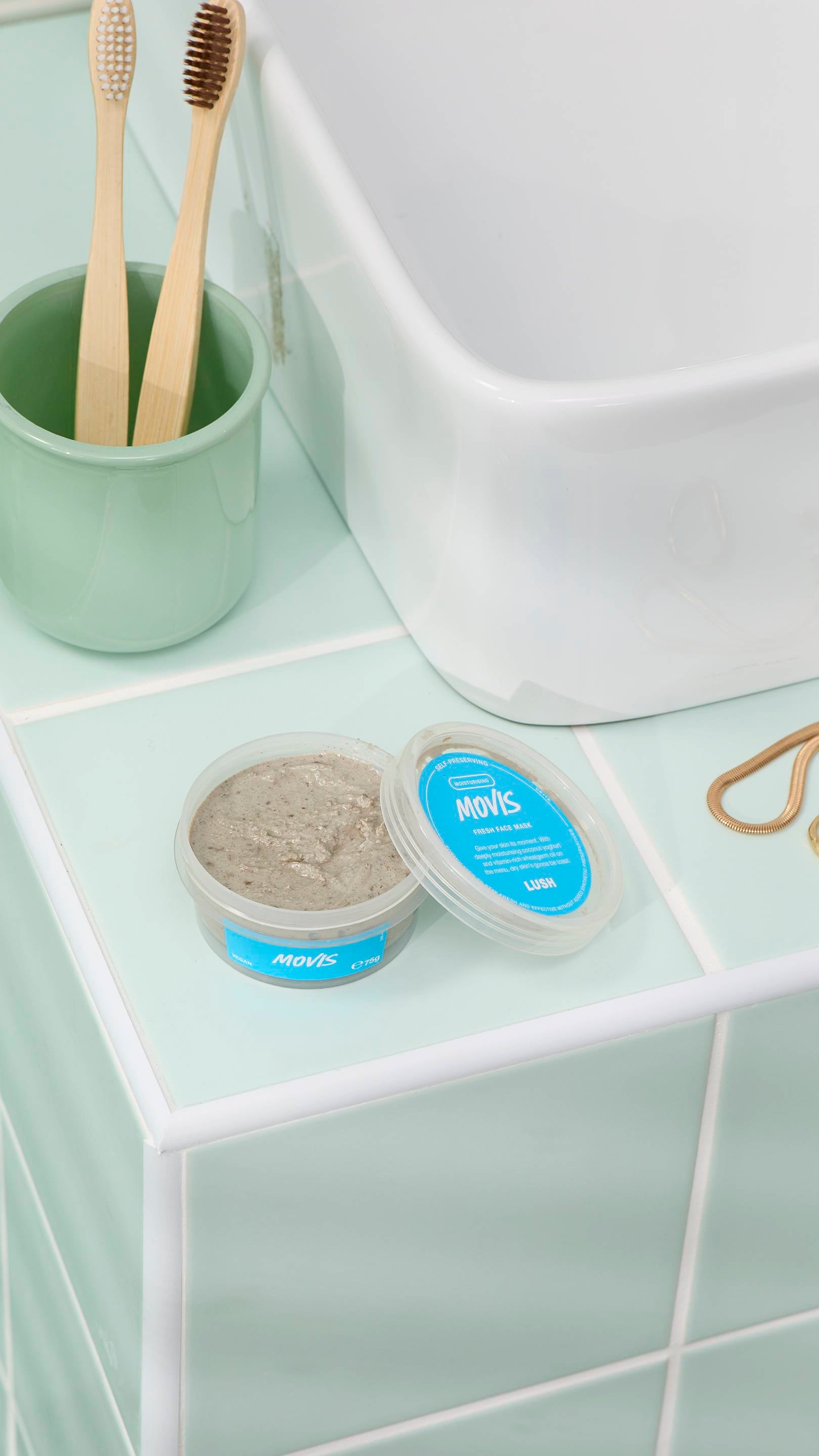 The image shows an open pot of grey, textured "Movis" face mask sitting on light green bathroom tiles next to a white sink. In the background, two bamboo toothbrushes are in a green cup. Gold jewellery rests nearby on the tiled surface.